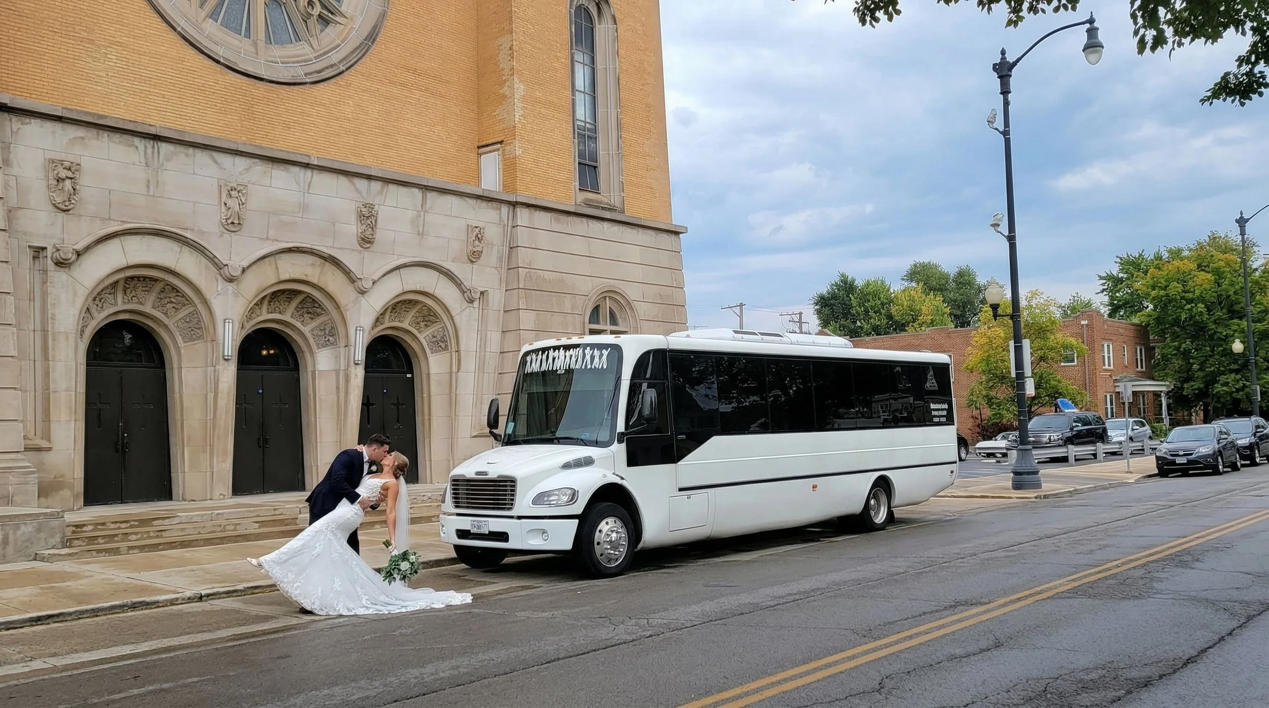 A bride and groom sharing a kiss outside a church on their wedding day, with a white shuttle bus parked beside them on the street.