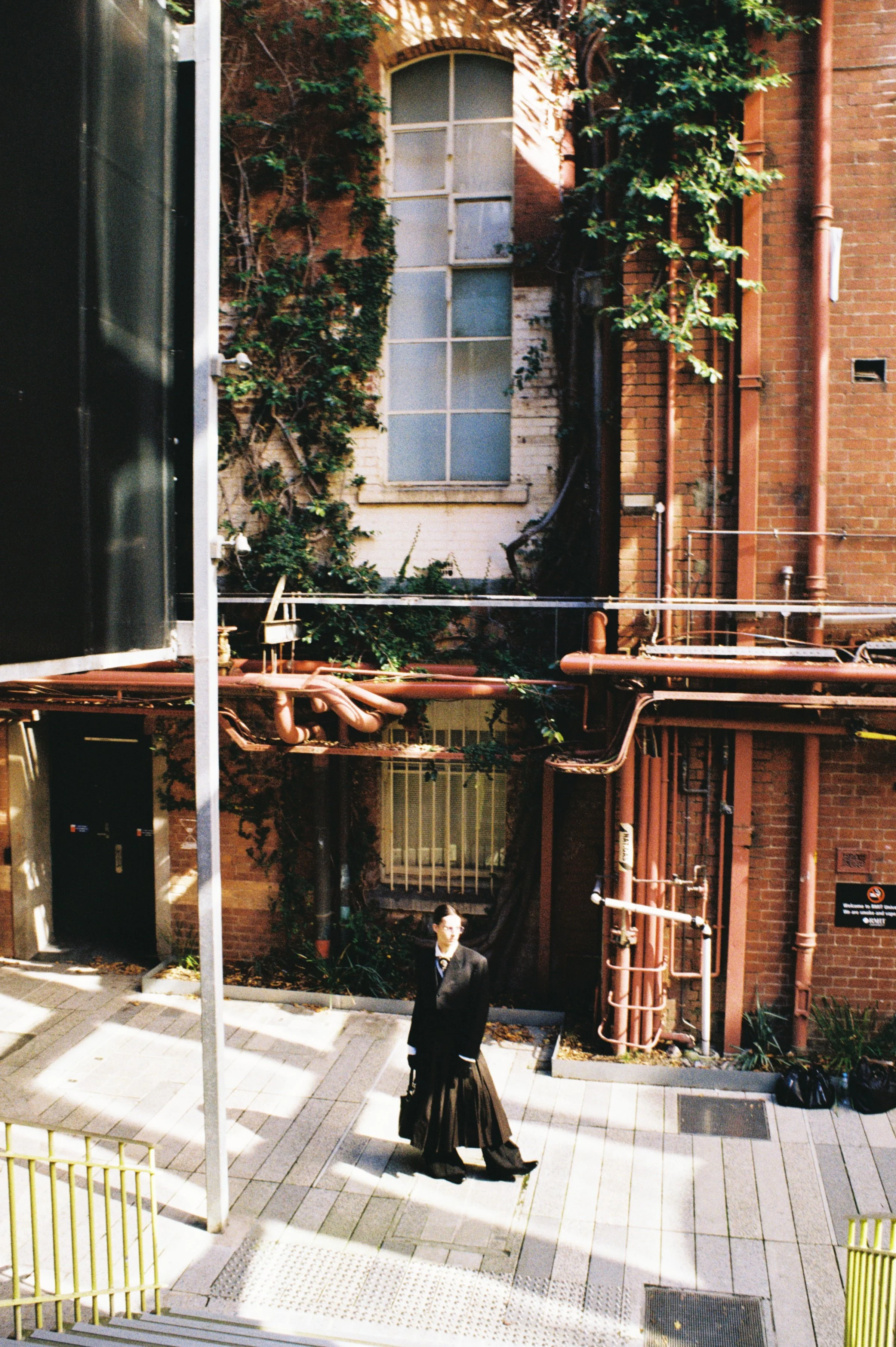 An urban alley with a red brick wall, metal pipes, a window, and a person in black robes carrying a bag standing on a paved sidewalk.