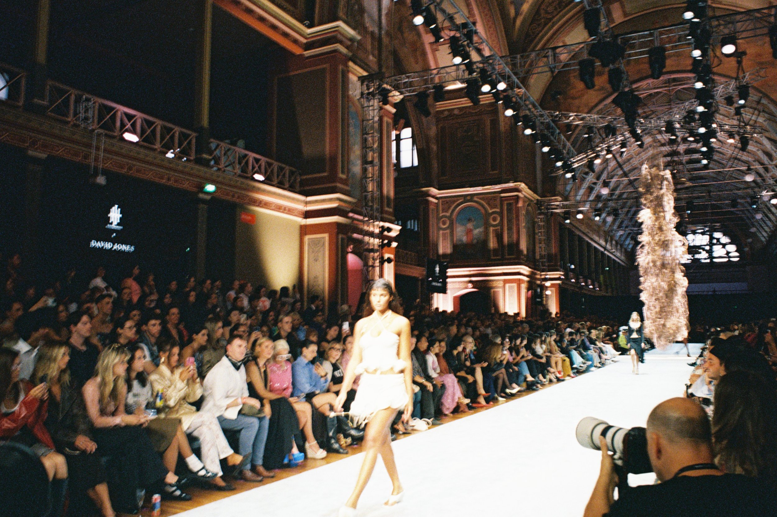 Fashion show event with a large seated audience in a historic, ornate hall, runway with a model walking, decorative wooden architecture, and professional lighting equipment overhead.