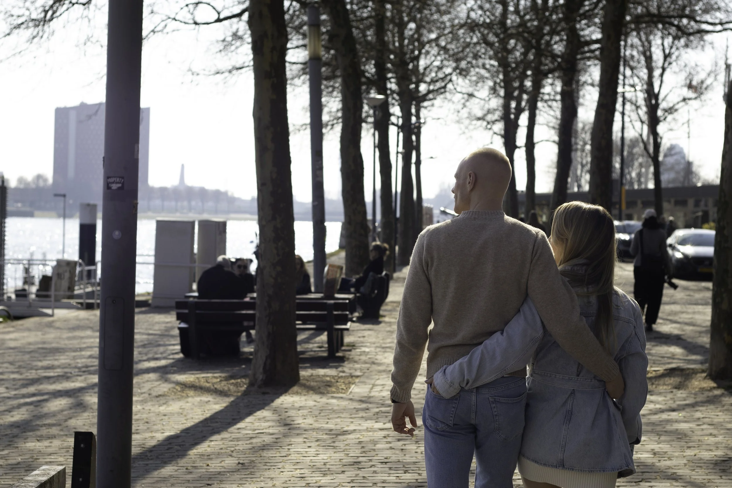 Man and woman with arms around each other, walking down a tree-lined path by a harbor
