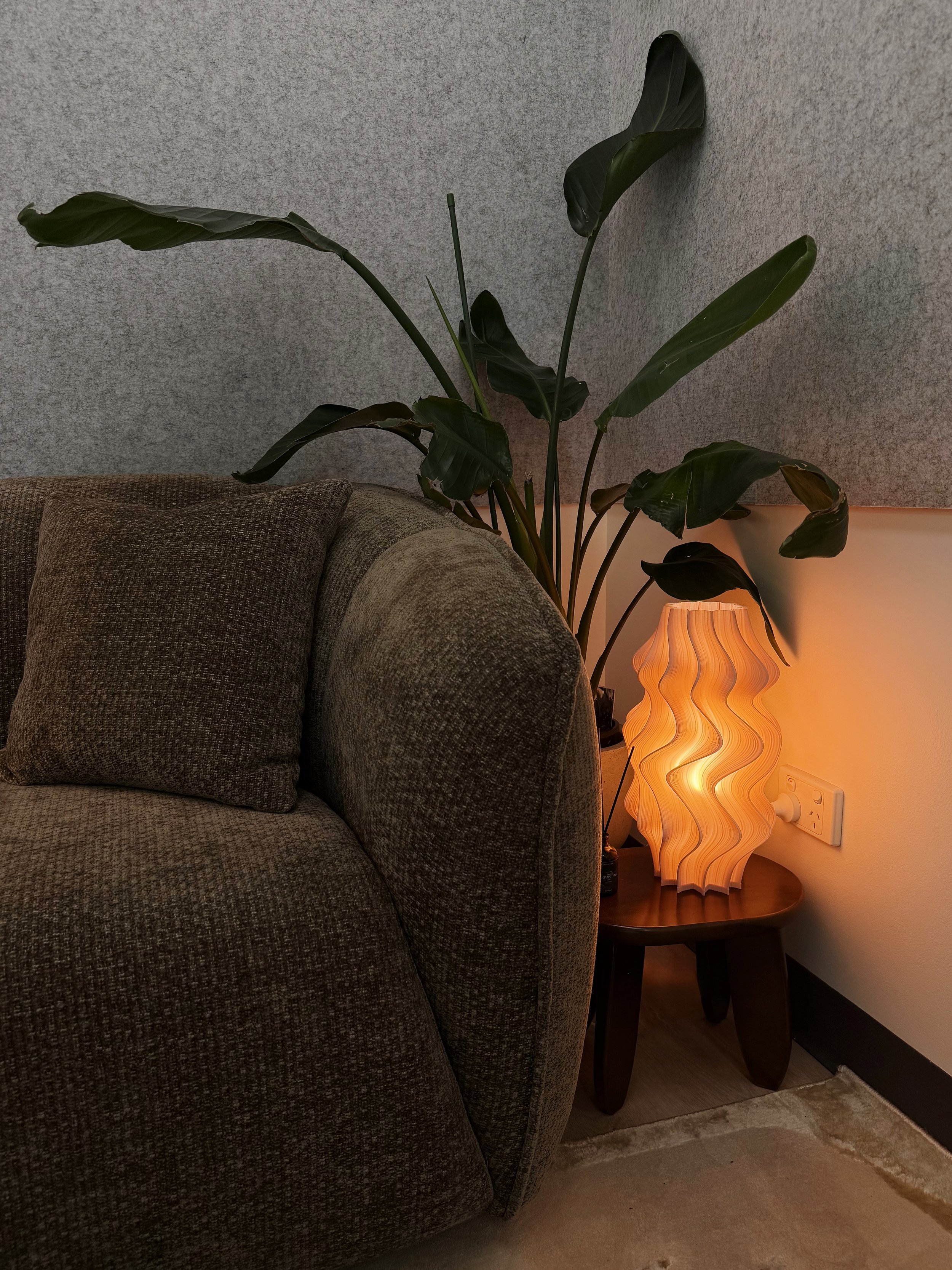 A cozy living room corner featuring a brown fabric sofa with matching pillows, a textured gray wall, a decorative table lamp with a wavy orange shade, and a tall green plant with large leaves.