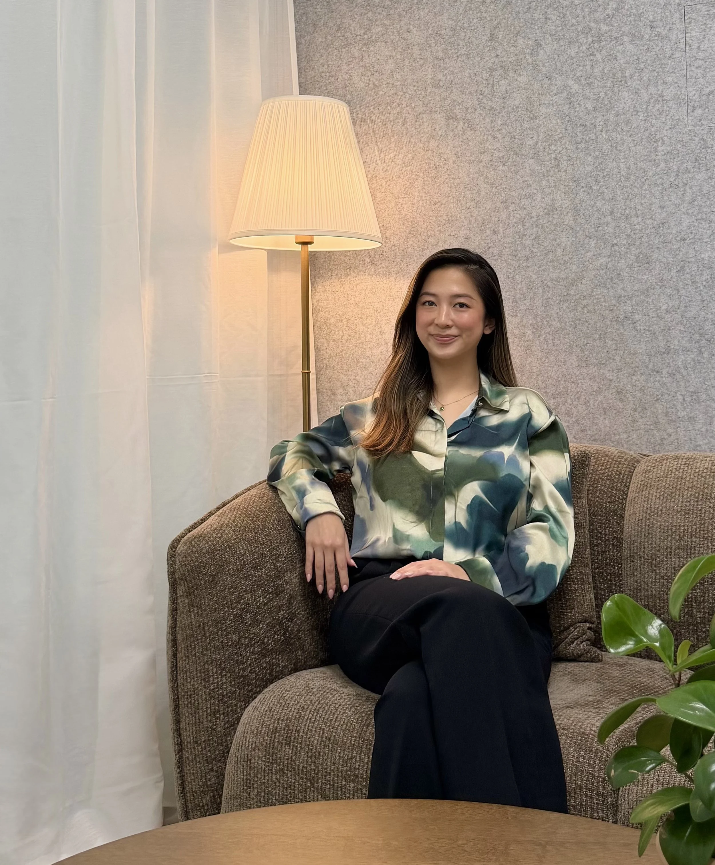 A young woman sitting on a brown upholstered chair, smiling, next to a modern floor lamp with a beige lampshade, in a room with gray textured wall, white curtains, and a green leafy plant in the foreground.