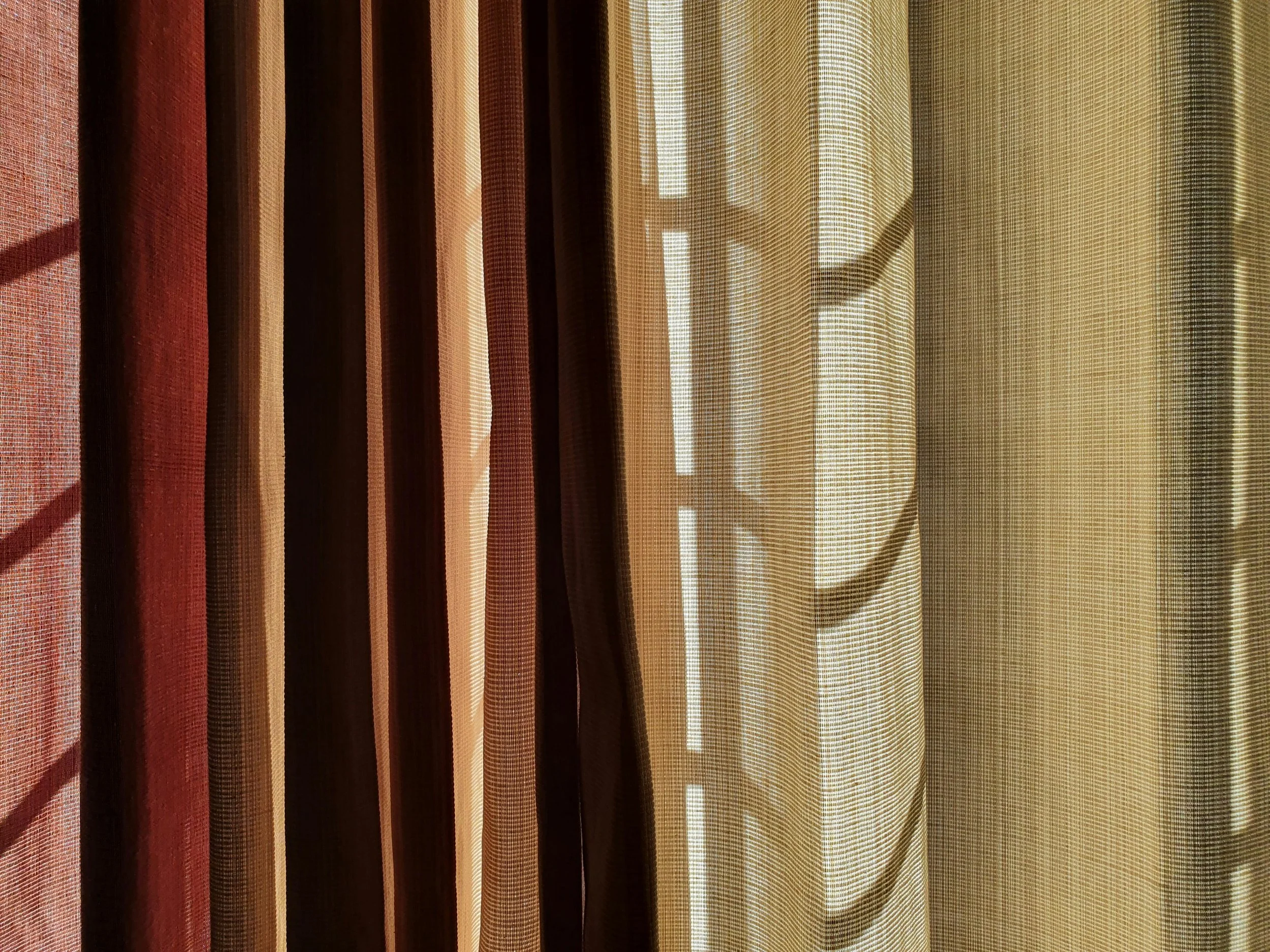A close-up of bus window curtains with alternating black, beige, and red vertical stripes, with sunlight coming through the fabric.
