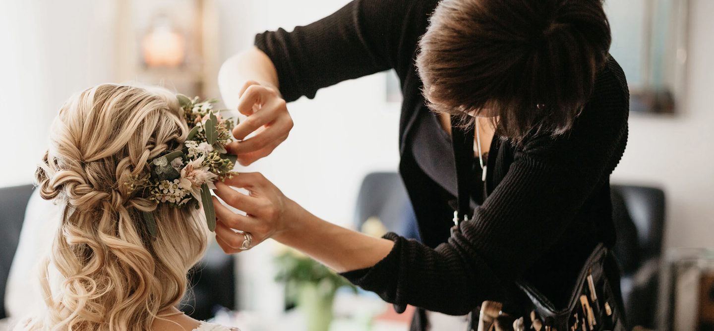 Maureen Zollinger placing a floral crown on a blonde woman's head, who has curly hair, in an indoor setting.
