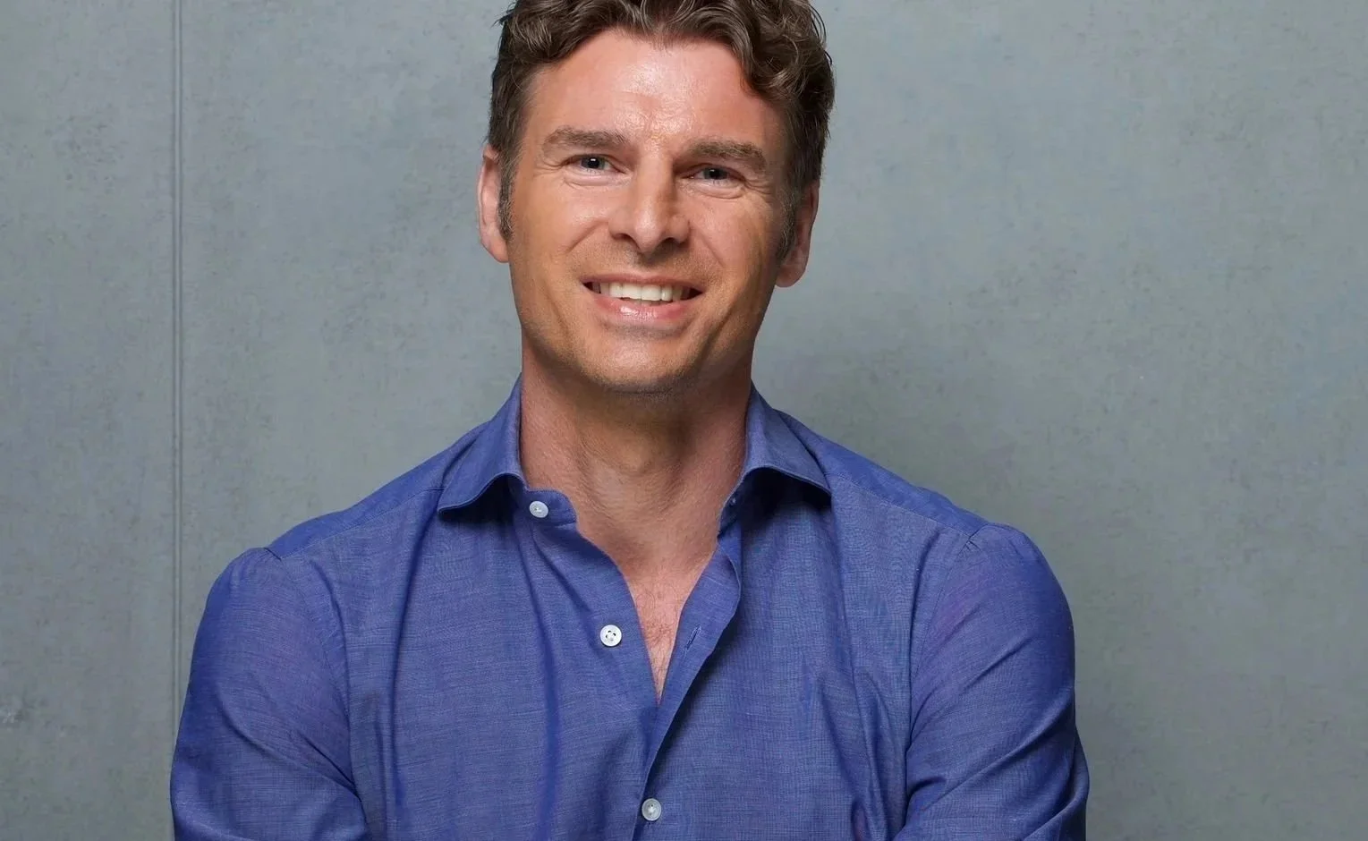 A smiling man wearing a blue dress shirt, standing against a plain grey background.