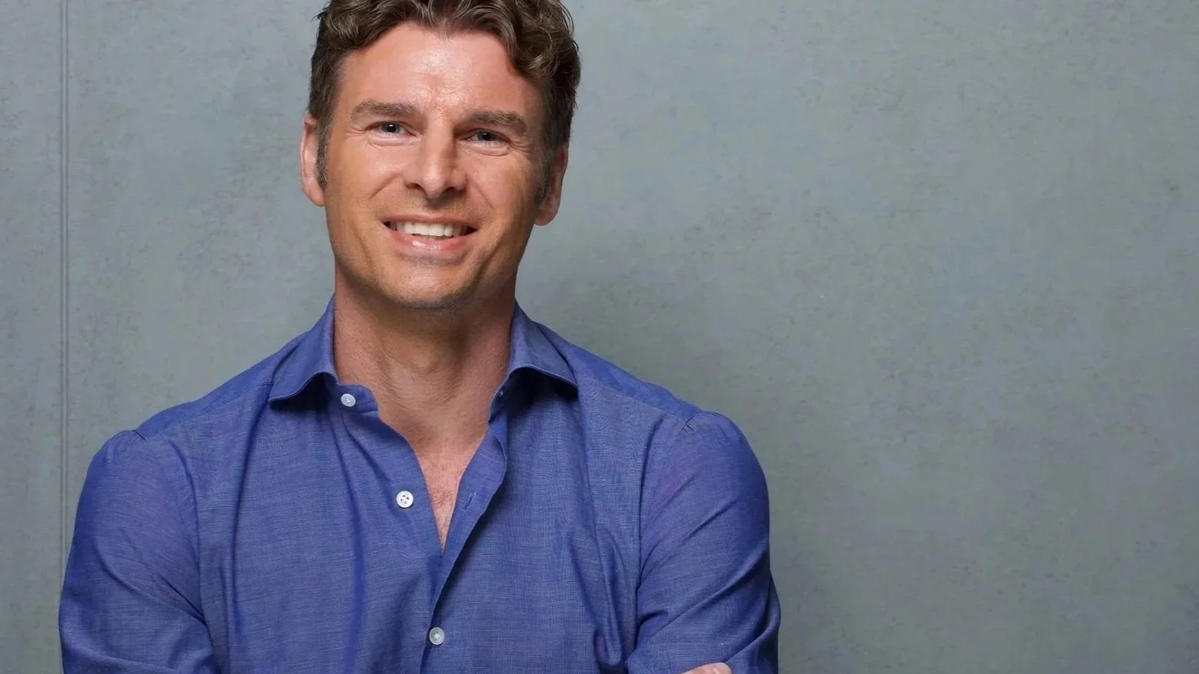 A man with brown hair and a smile wearing a blue button-up shirt standing in front of a plain gray wall.