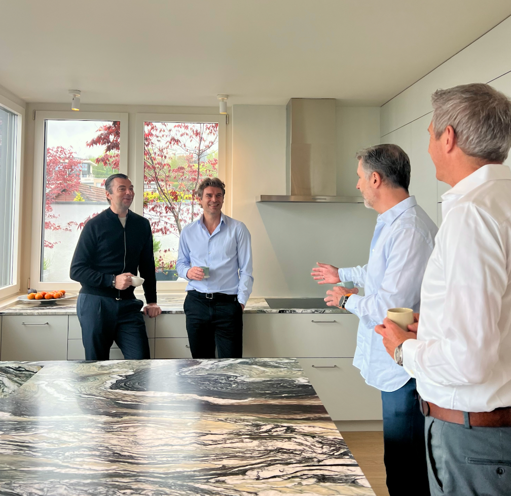 Four men having a conversation and drinking coffee in a modern kitchen with large windows showing trees outside.