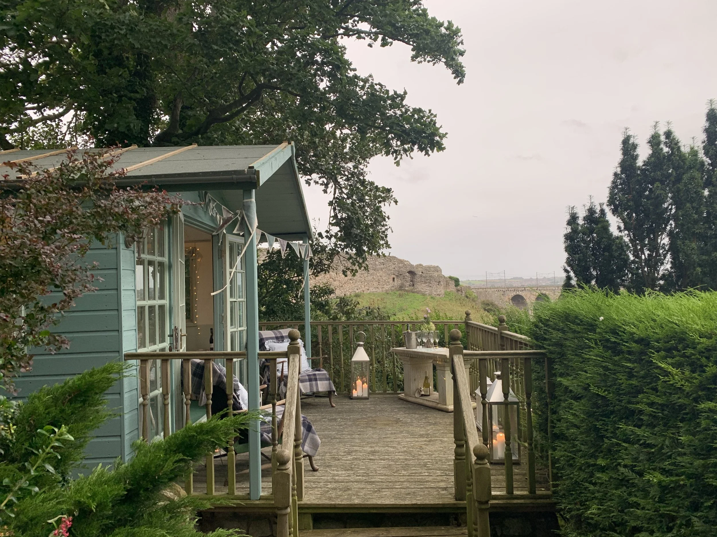 Sage-green summerhouse deck decorated with bunting and lanterns styled by Laura Somborn, surrounded by garden greenery and overlooking the ruins of Berwick Castle,