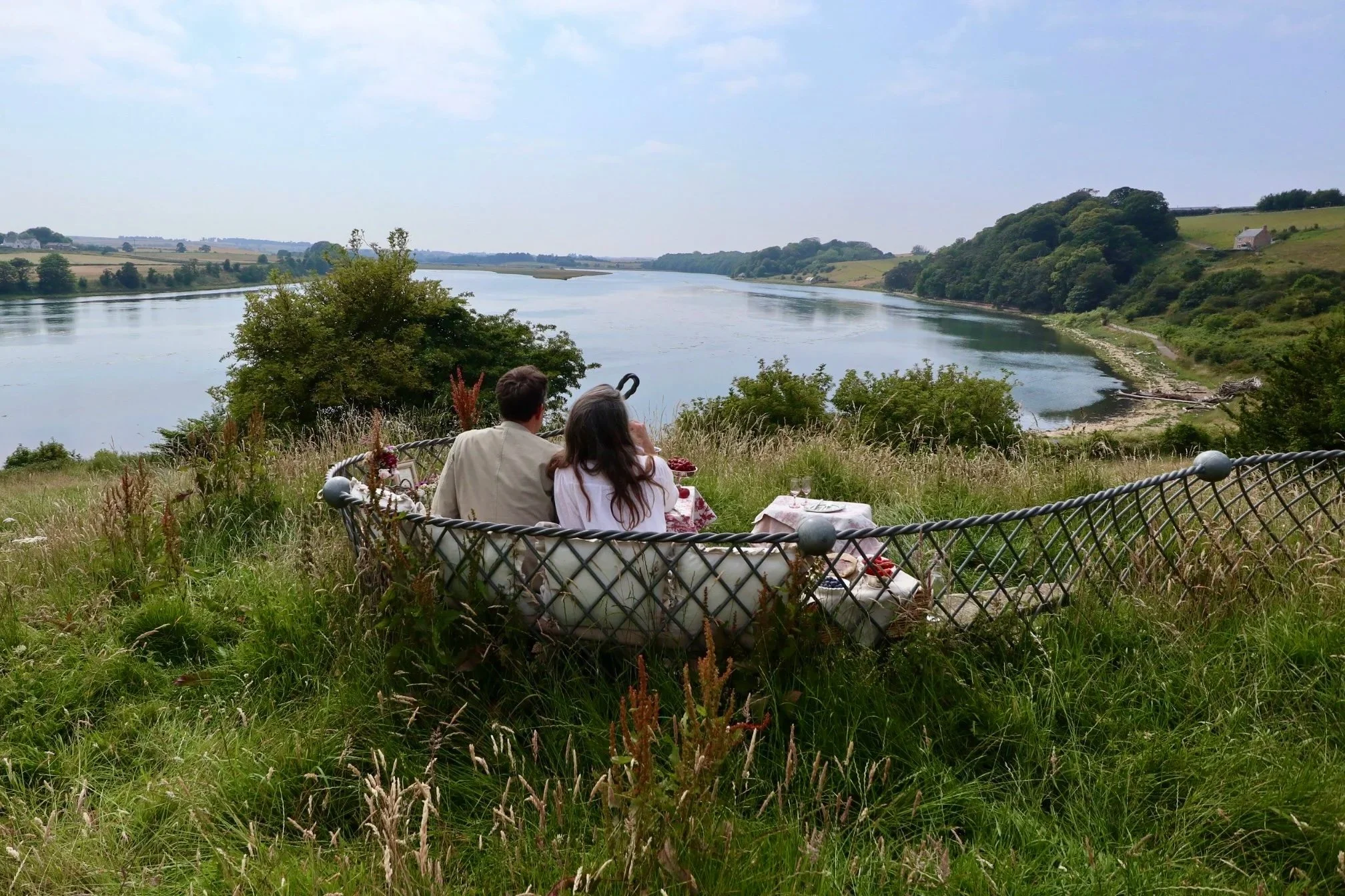 A couple sitting on a vintage sofa outdoors overlooking a river and green hills, accompanying a picnic setup with food on a table.