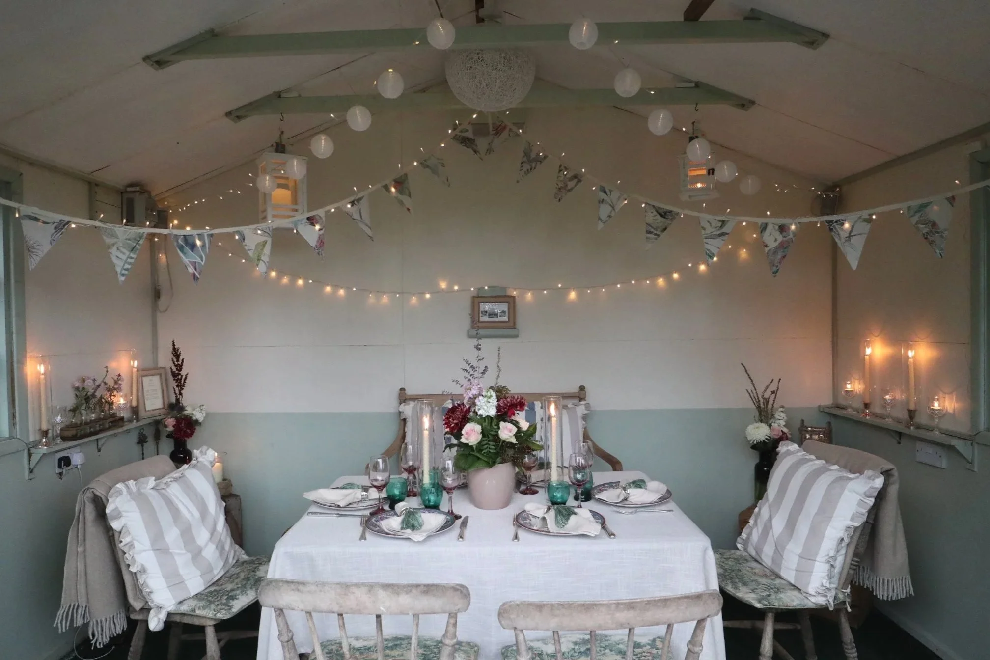 Intimate summerhouse tablescape styled by Laura Somborn with heritage tableware, seasonal floral arrangement, striped cushions and warm fairy lights.