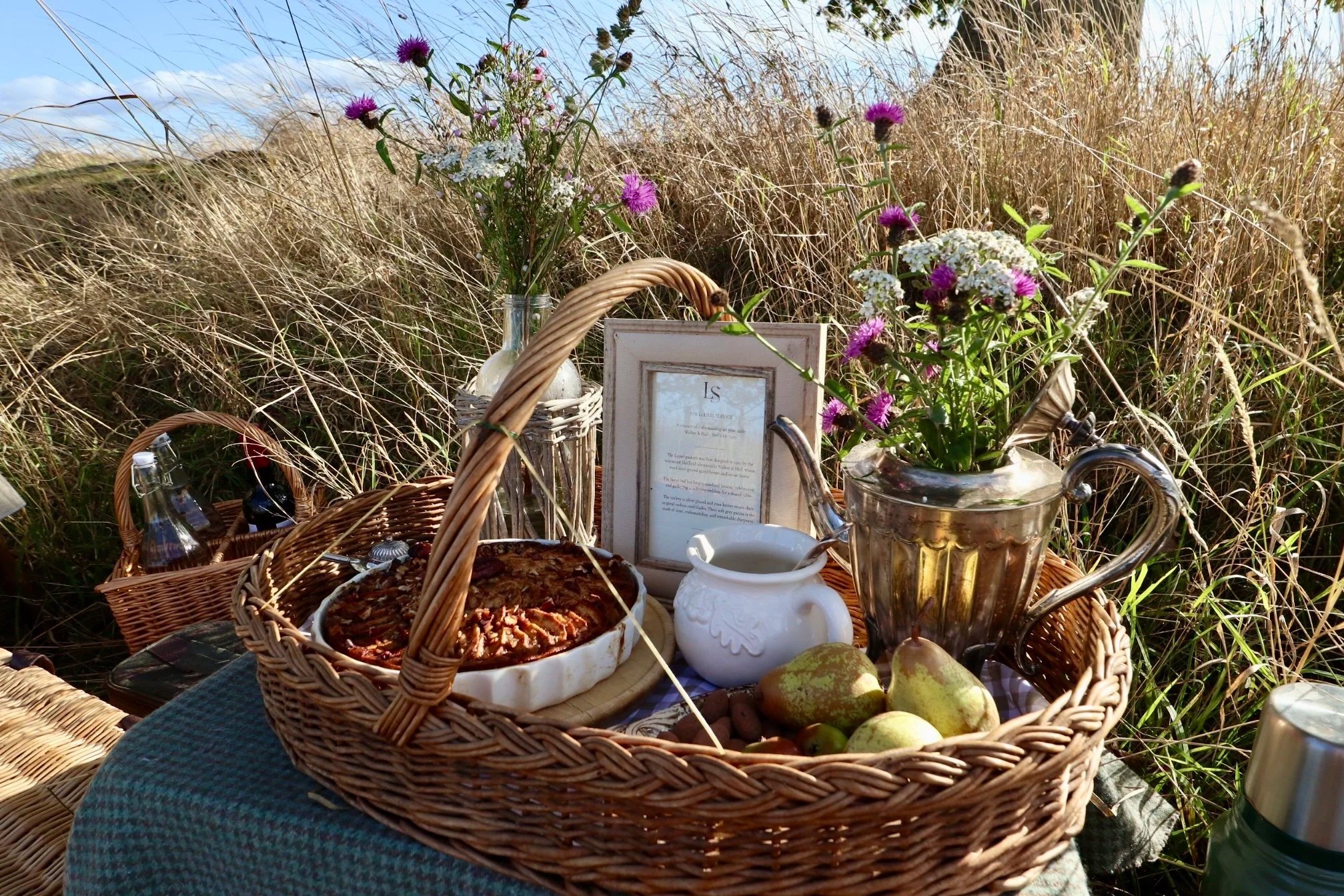 Wicker basket with fruit cake, pears and vintage serving pieces, arranged for a country gathering.