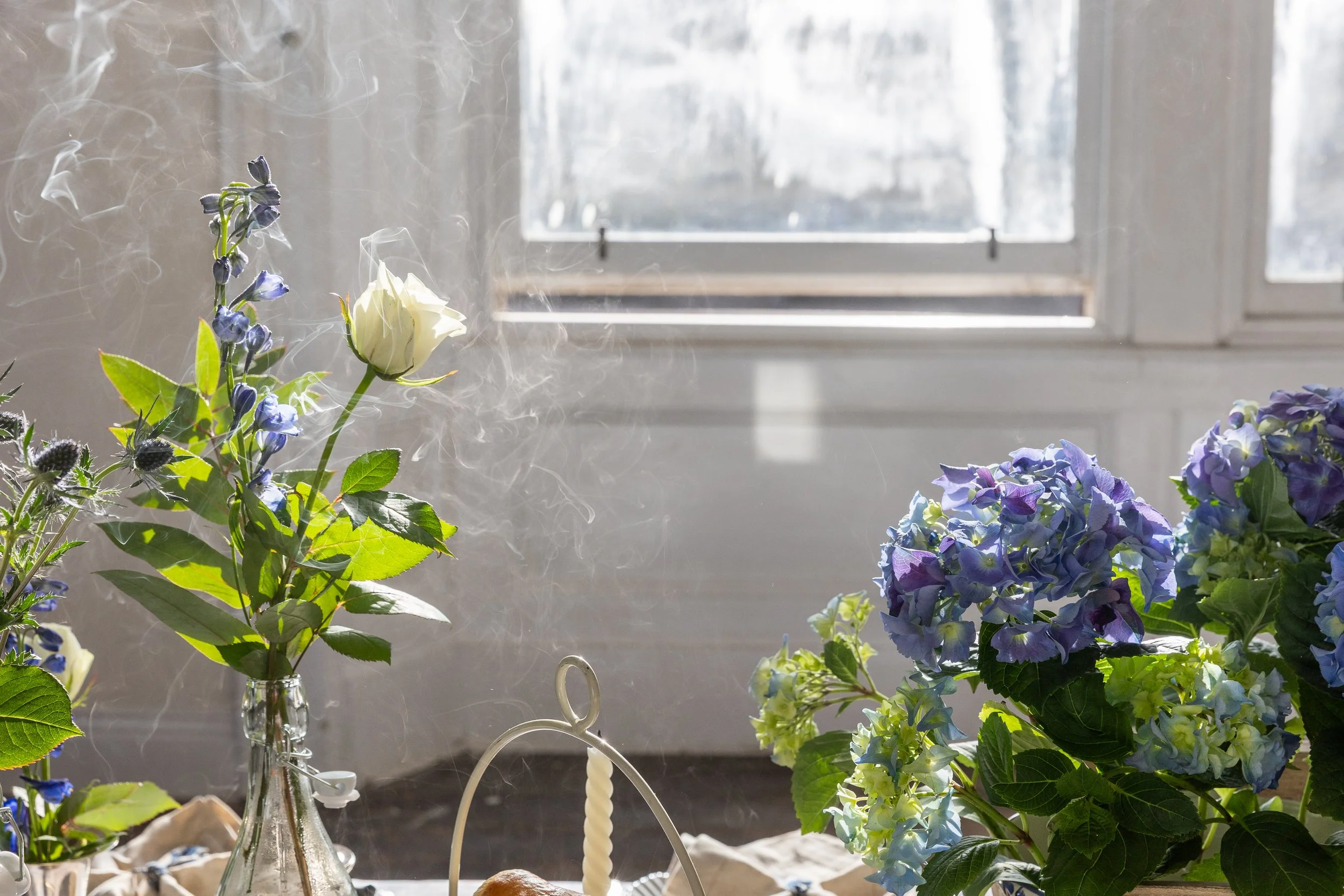 Atmospheric table setting with blue and white flower arrangement and blue hydrangeas in a sunlight room with sash windows.