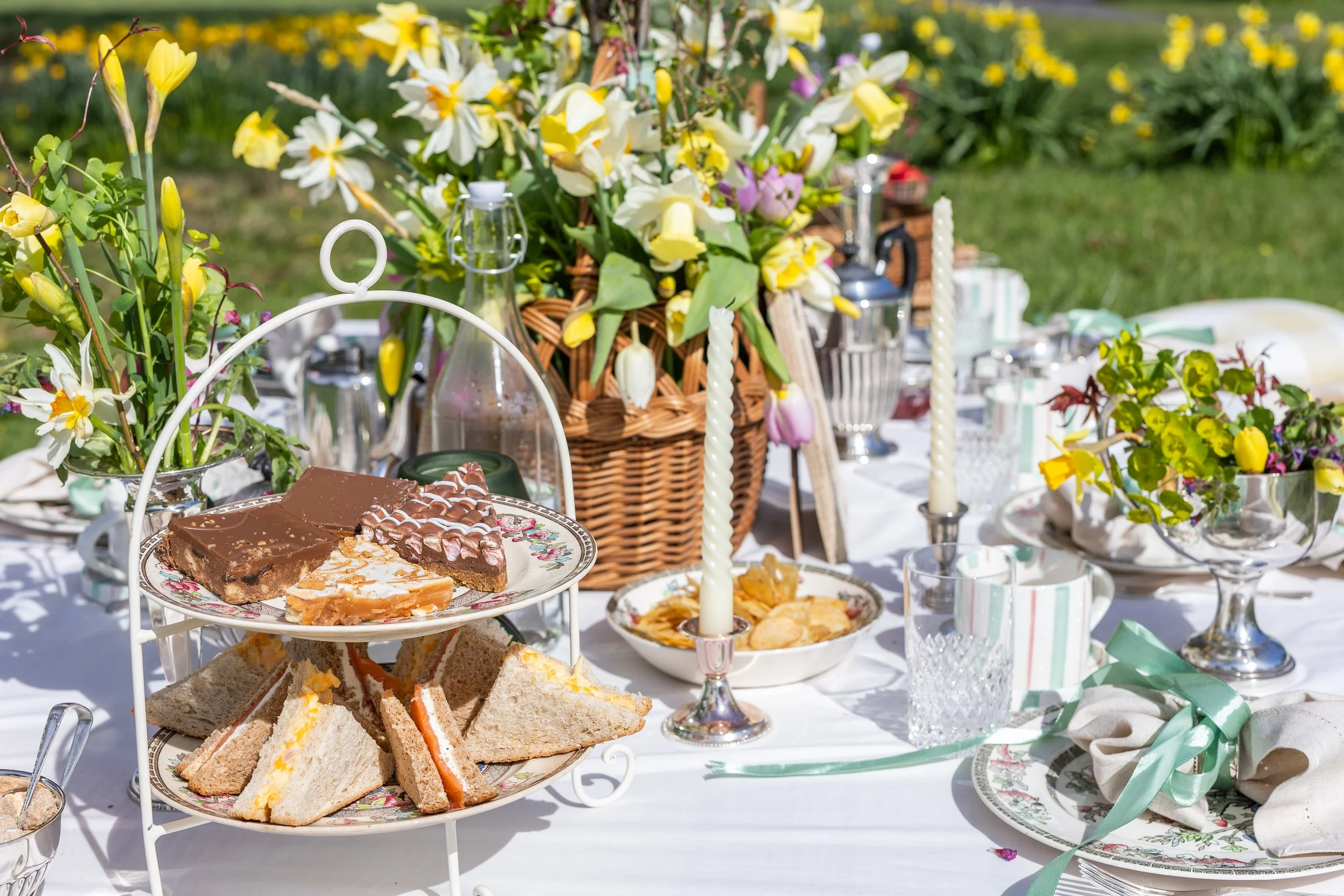 Outdoor picnic gathering by Laura Somborn with floral arrangements, set for afternoon tea with silver cutlery, linen napkins and softly layered table details.