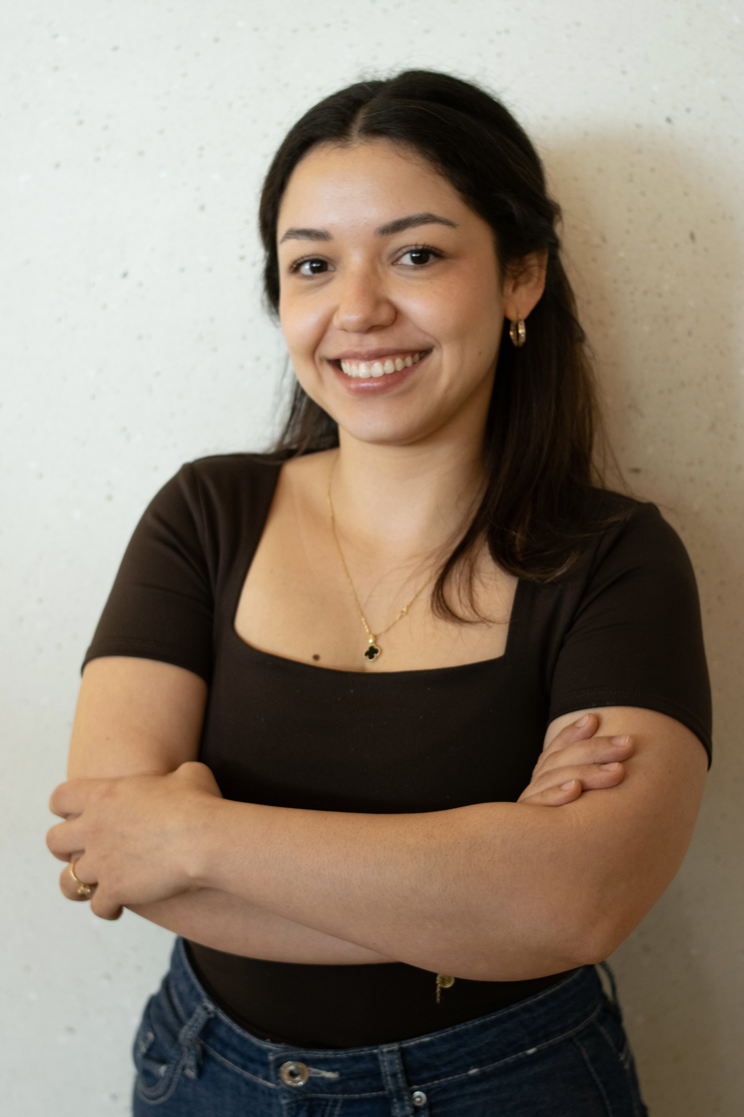 Mujer joven con cabello oscuro, usando un top negro y jeans, sonriendo con los brazos cruzados, frente a una pared beige.