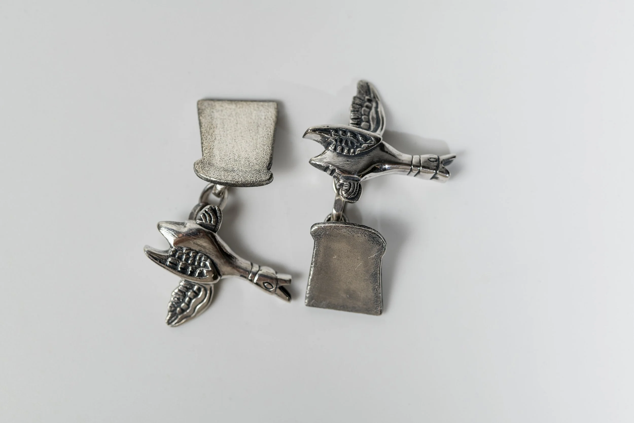 Silver cufflinks with hand carved ducks on one side and a slice of bread on the other, arranged on a white surface.