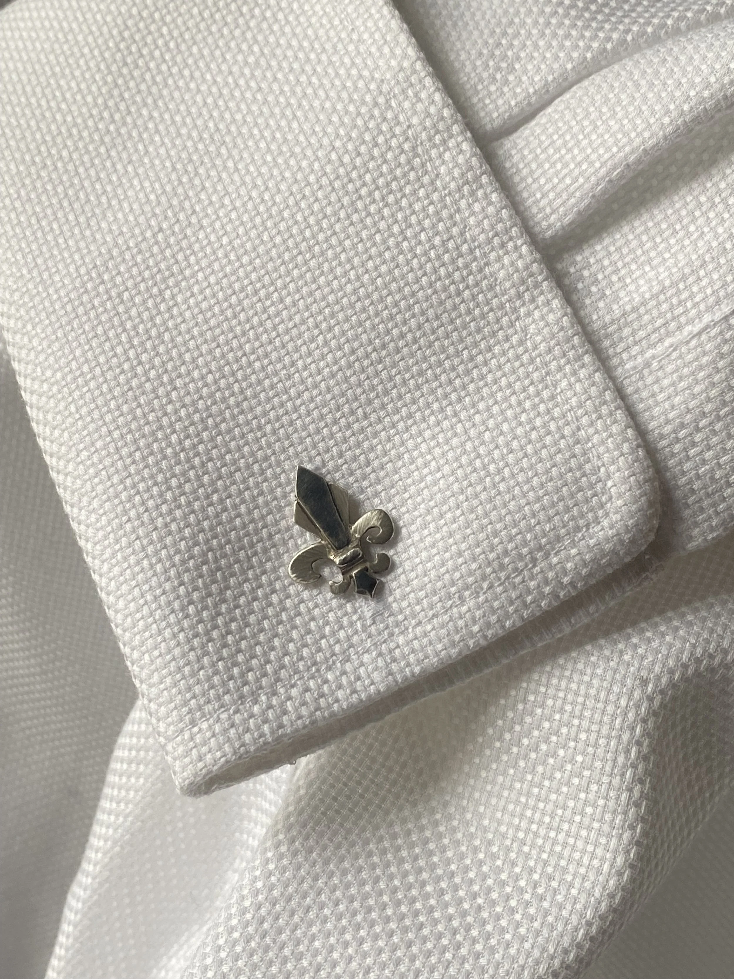 A silver Fleur-de-lis cufflink on white textured dress shirt cuff.