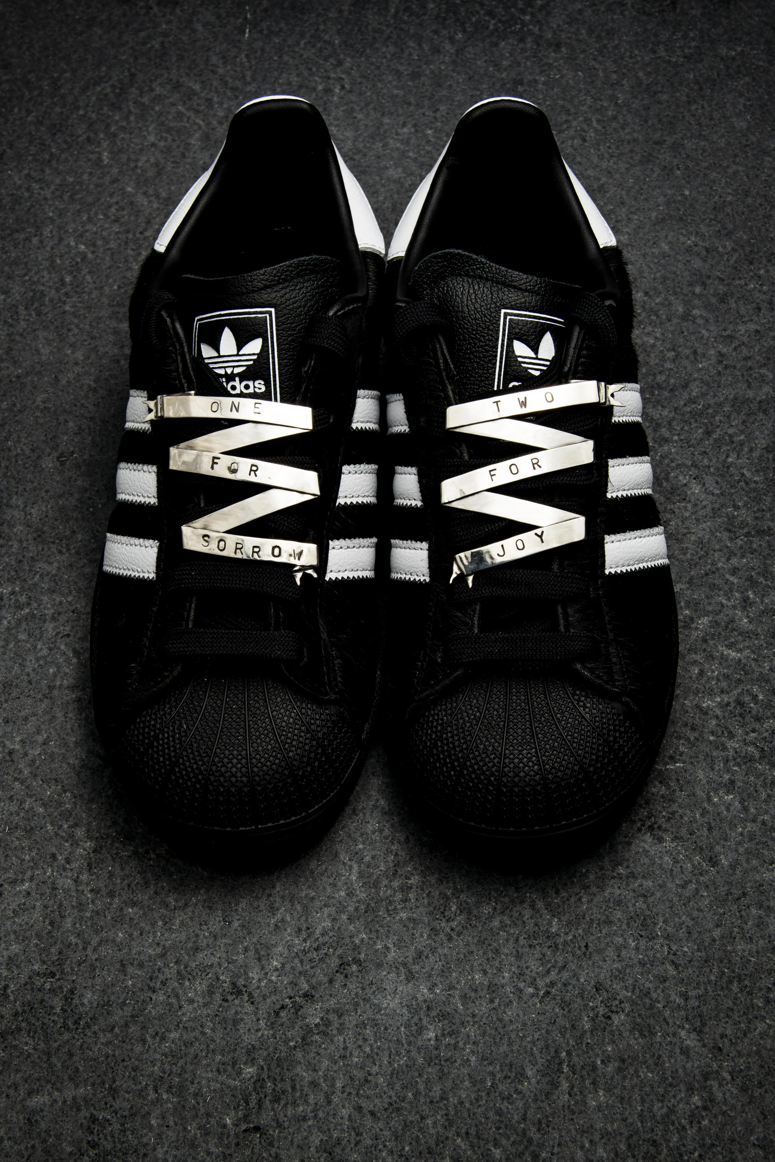 Pair of black and white Adidas sneakers on a dark textured surface with decorative laces charm ribbon that reads "One for sorrow," "Two for joy."