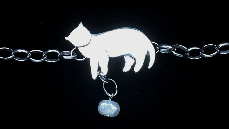 A silver bracelet with a charm of a cat hanging upside down from a chain, holding a small pearl-shaped pendant.