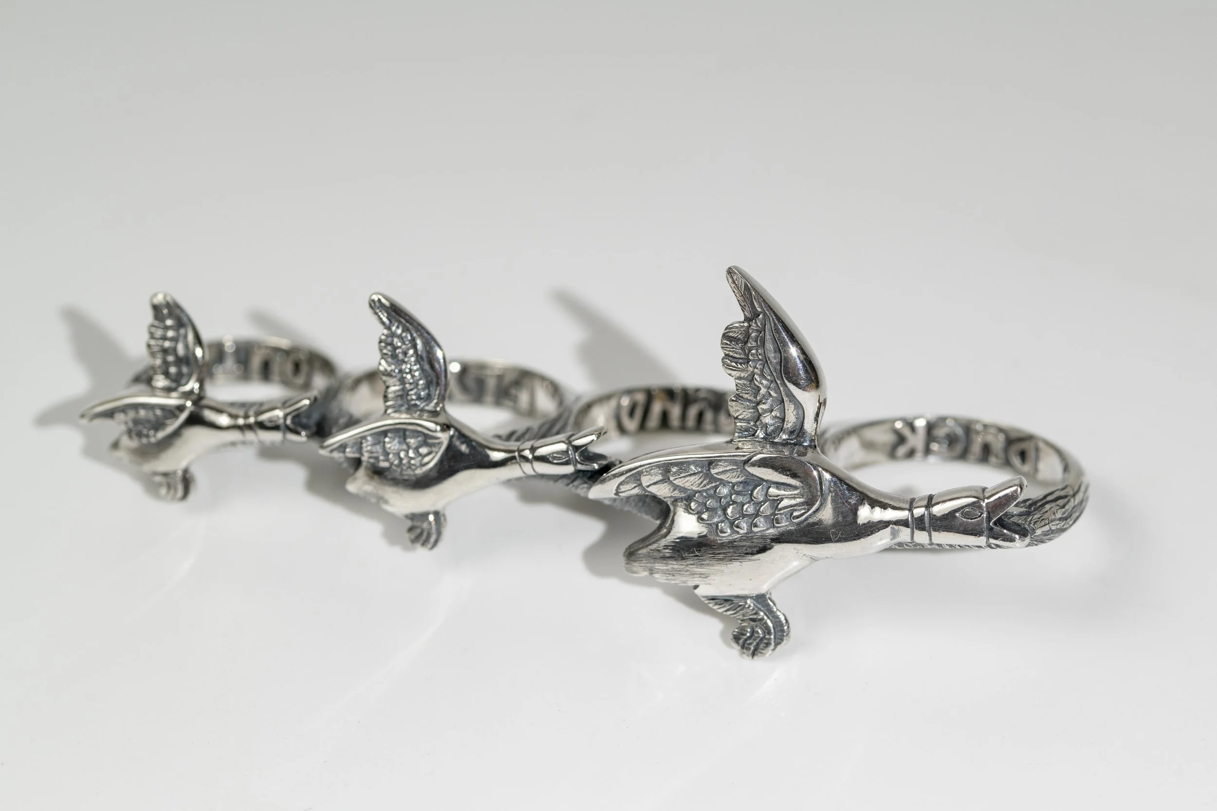 Sterling Silver 4-finger ring in the shape of three flying ducks all graduating in size, with detailed feathers. The engraving of 'Duck Around Fly Out' on the inside of the fingers. Lying on a gloss white surface.