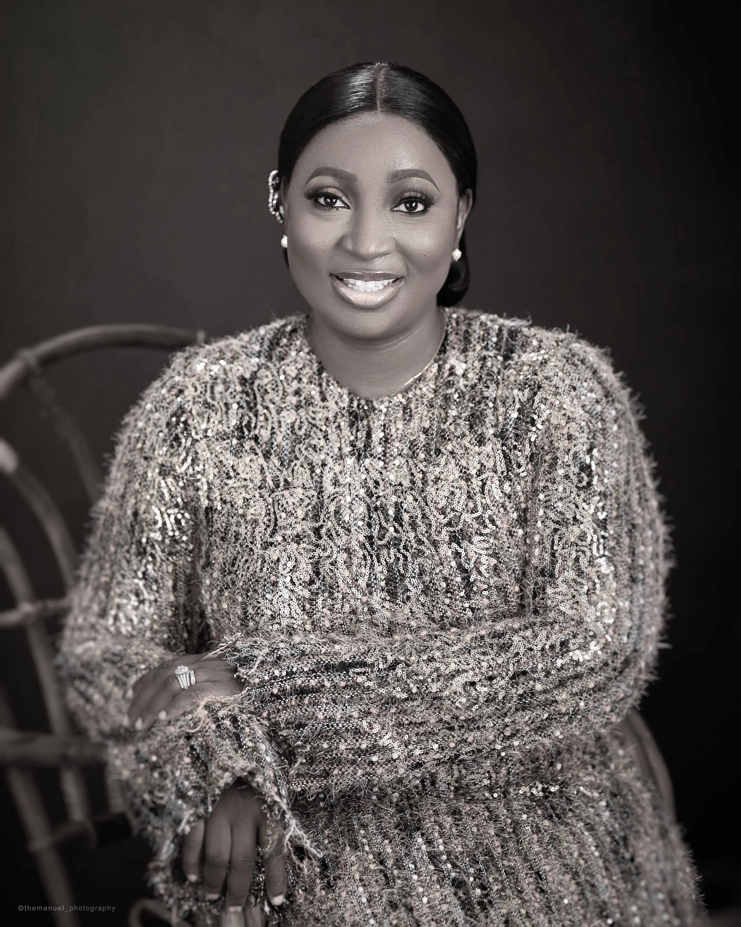 A close-up black and white portrait of a woman smiling, wearing an ornate, sparkling dress and pearl earrings, with a seated pose and her evn-hand displaying a ring.