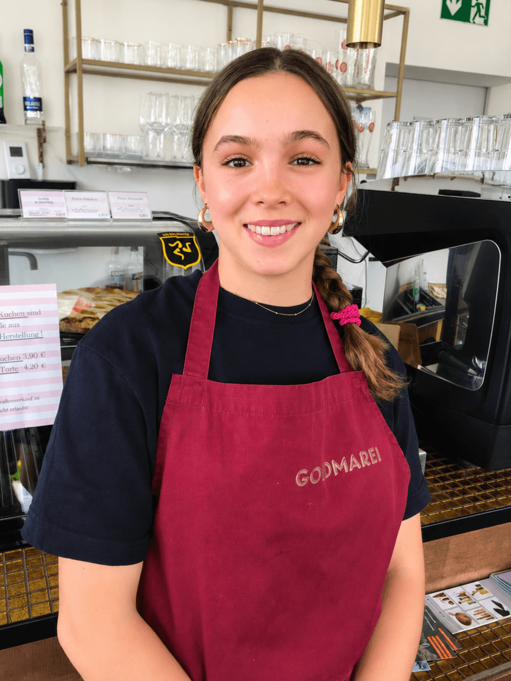 Lächelnde junge Frau mit langen braunen Haaren in Zopf, trägt eine schwarze T-Shirt und rote Schürze mit Aufschrift 'GOOMAREI', steht hinter einem Glastresen in einem Café oder einer Bäckerei.