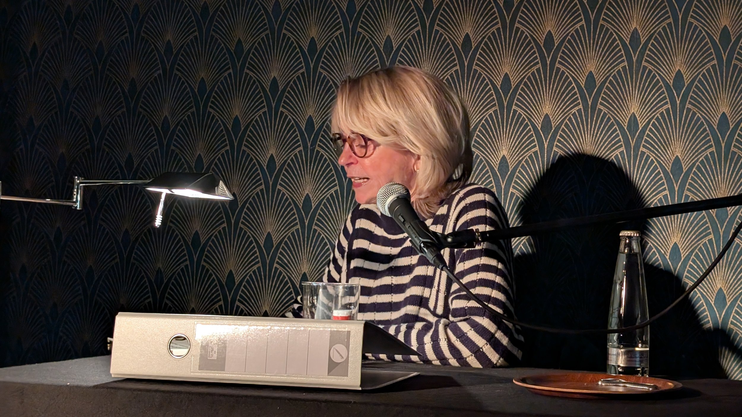 Frau mit Brille spricht in ein Mikrofon bei einem Tisch, im Hintergrund eine tropfenförmige Musterwand, auf dem Tisch liegen Wasserflasche, Glas und eine Box.