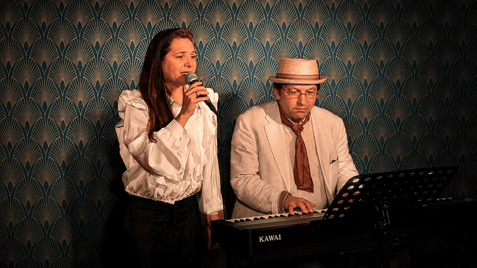 Zwei Musiker, eine Frau mit Mikrofon in der Hand und ein Mann an einem Keyboard, vor einer kunstvollen Tapeten