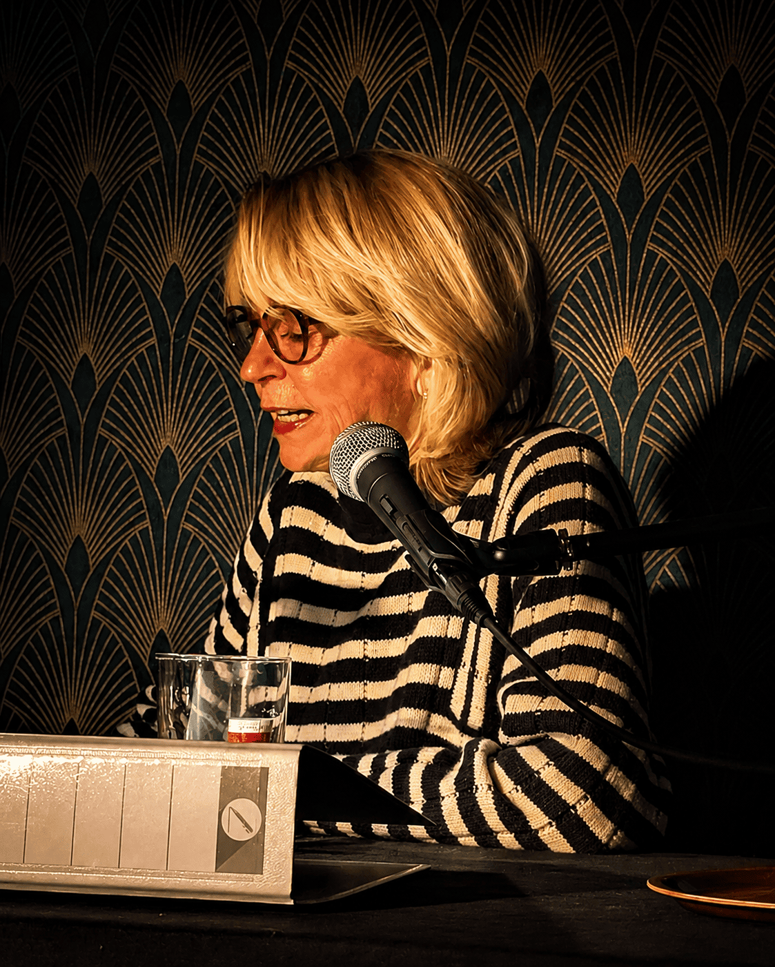 Eine Frau mit blonden Haaren und Brille sitzt an einem Tisch, spricht in ein Mikrofon, trägt ein schwarz-weiß gestreiftes Oberteil, und hat ein Glas Wasser vor sich. Die Wand im Hintergrund hat ein dekoratives Muster.