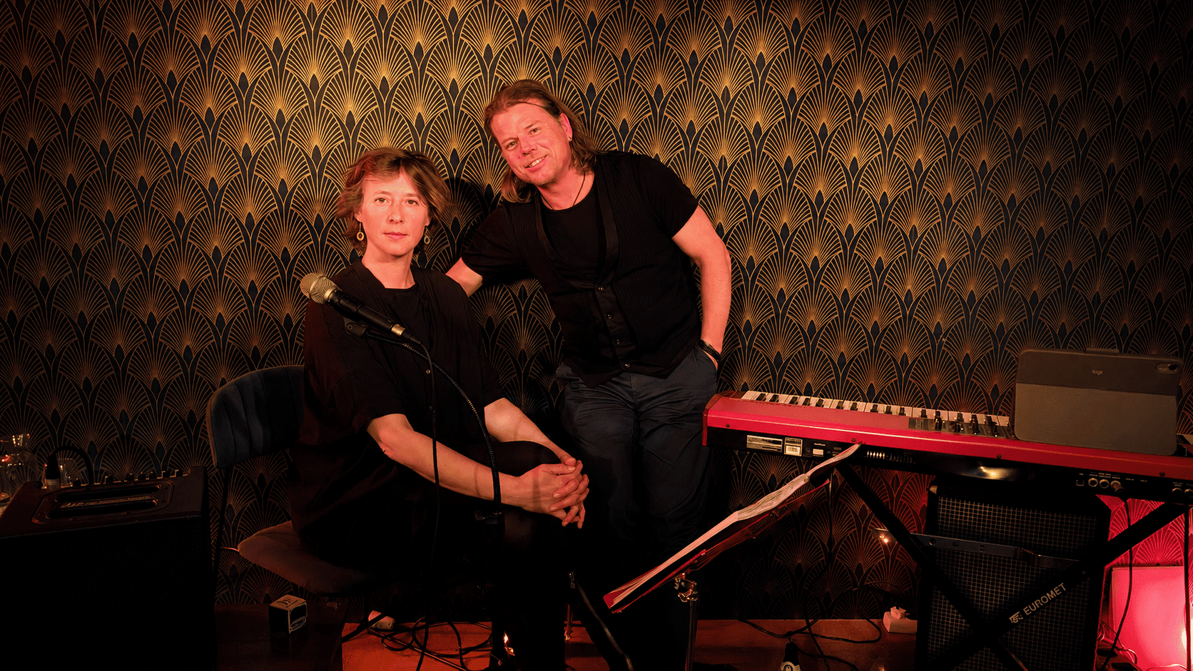 Ein Mann und eine Frau in einem Musikraum mit dunkler, goldverzierter Wand hinter ihnen, umgeben von Musikinstrumenten wie einem Keyboard, einem Mikrofon und einem Verstärker.