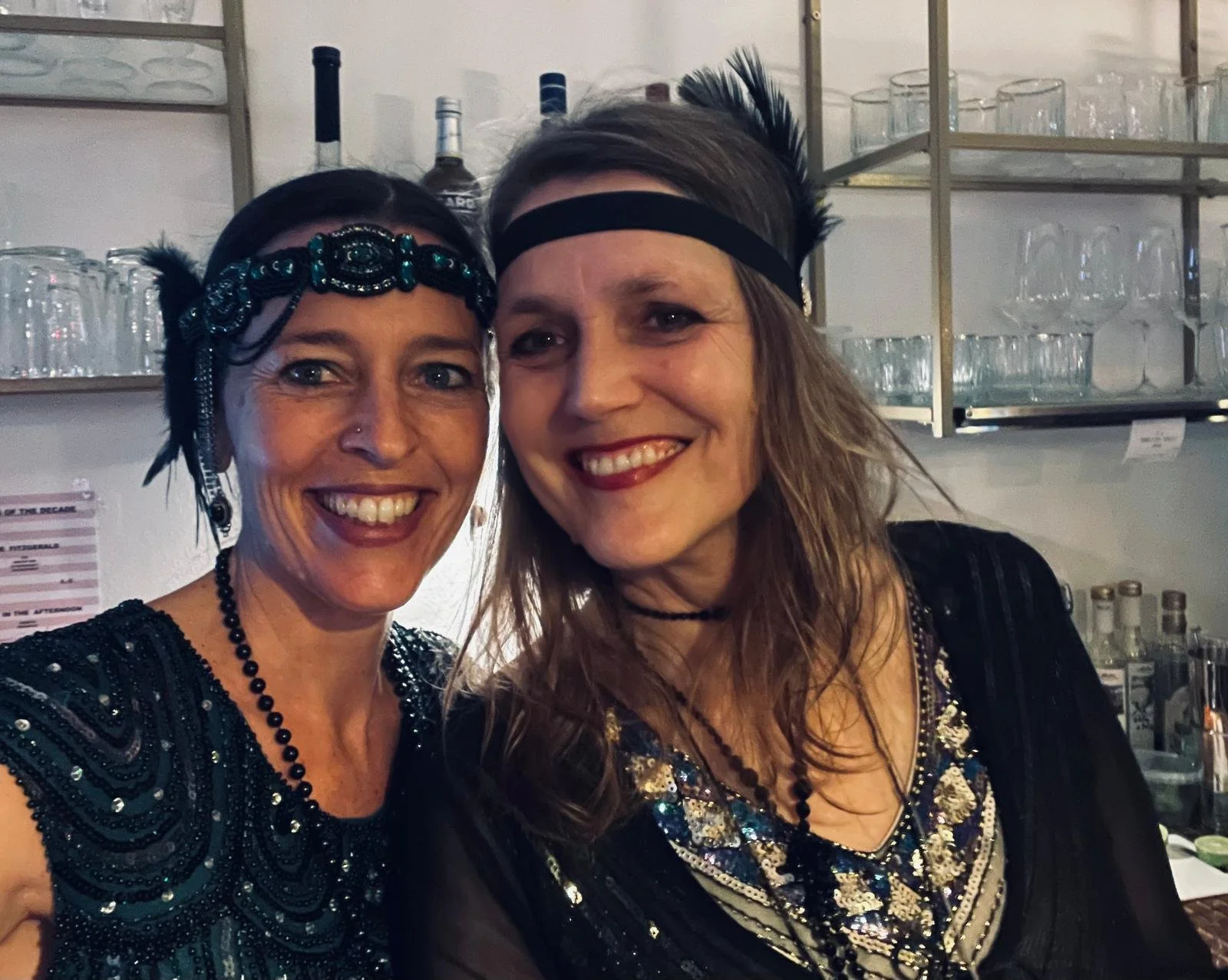 Zwei Frauen in Silvesterkostümen posieren für ein Selfie im Barbereich, im Hintergrund sind Gläser und Flaschen sichtbar.