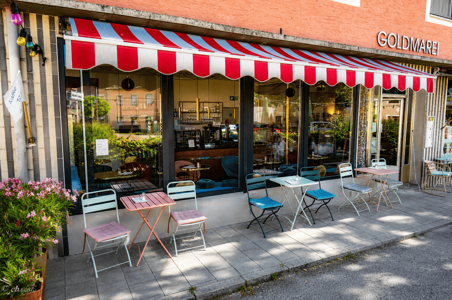 Außenansicht eines Cafés mit Tischen und Stühlen auf der Terasse, einer roten und weißen Markise, Blumen im Vordergrund und einem Blick ins Innere des Cafés, das mit modernen Möbeln und Dekorationen ausgestattet ist.