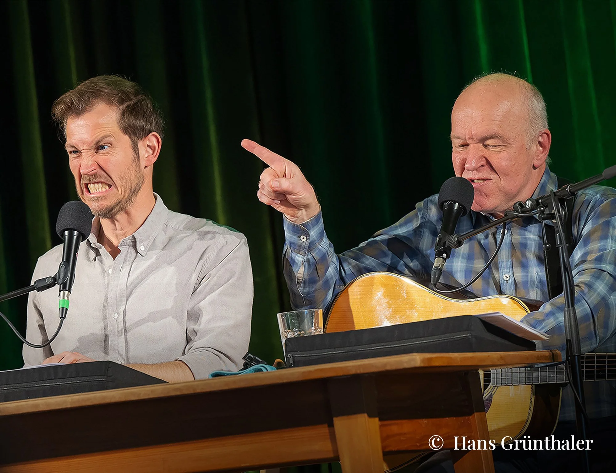 Zwei Männer sitzen an einem Tisch, der eine spielt Gitarre und singt in ein Mikrofon, der andere sieht ihn mit einem ausdrucksstarken Gesicht an. Hintergrund mit grünen Vorhängen.