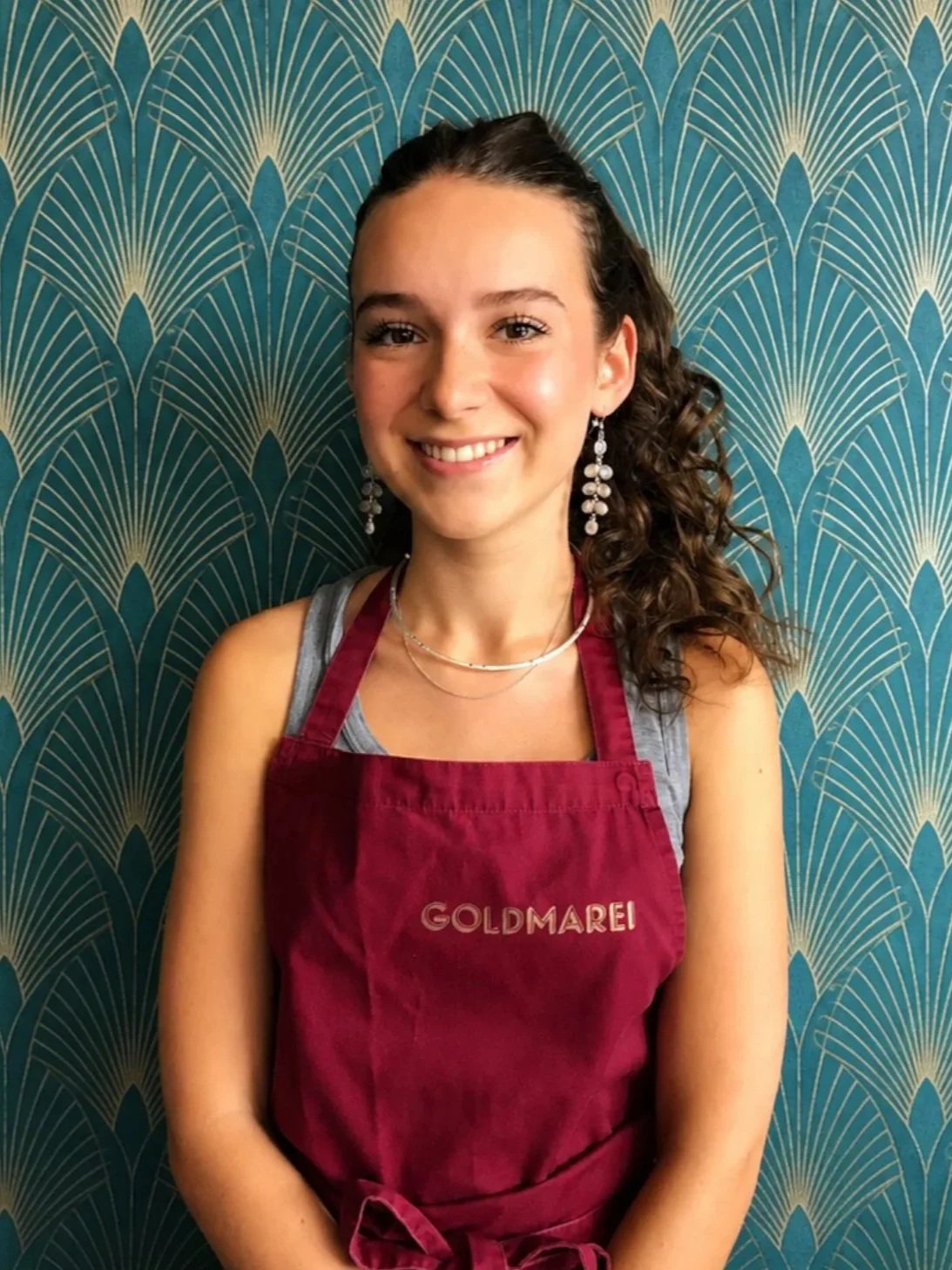 Junge Frau trägt eine braune Schürze mit 'GOLDMAREI' Aufdruck, lächelt, hat dunkle, lockige Haare, trägt Ohrringe und Ketten, vor einer blauen, geometrischen Tapete.