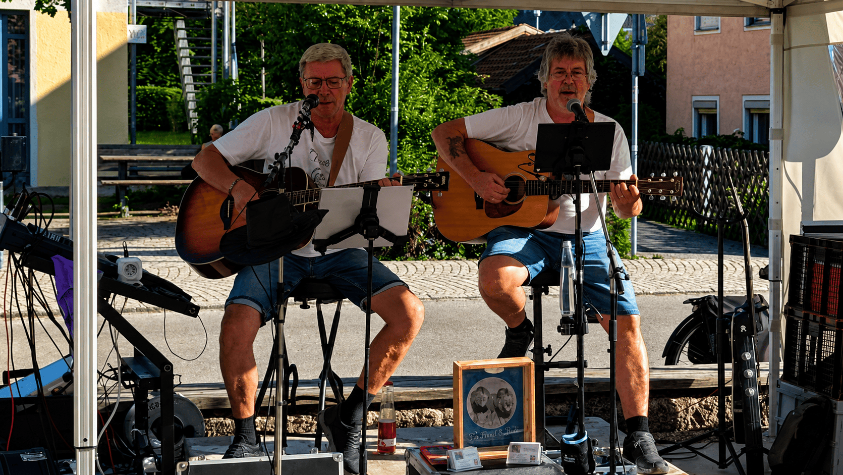 Zwei Männer spielen Gitarre auf einer Bühne im Freien, während sie singen und Musik machen, umgeben von Straßen, Häusern und Bäumen bei sonnigem Wetter.