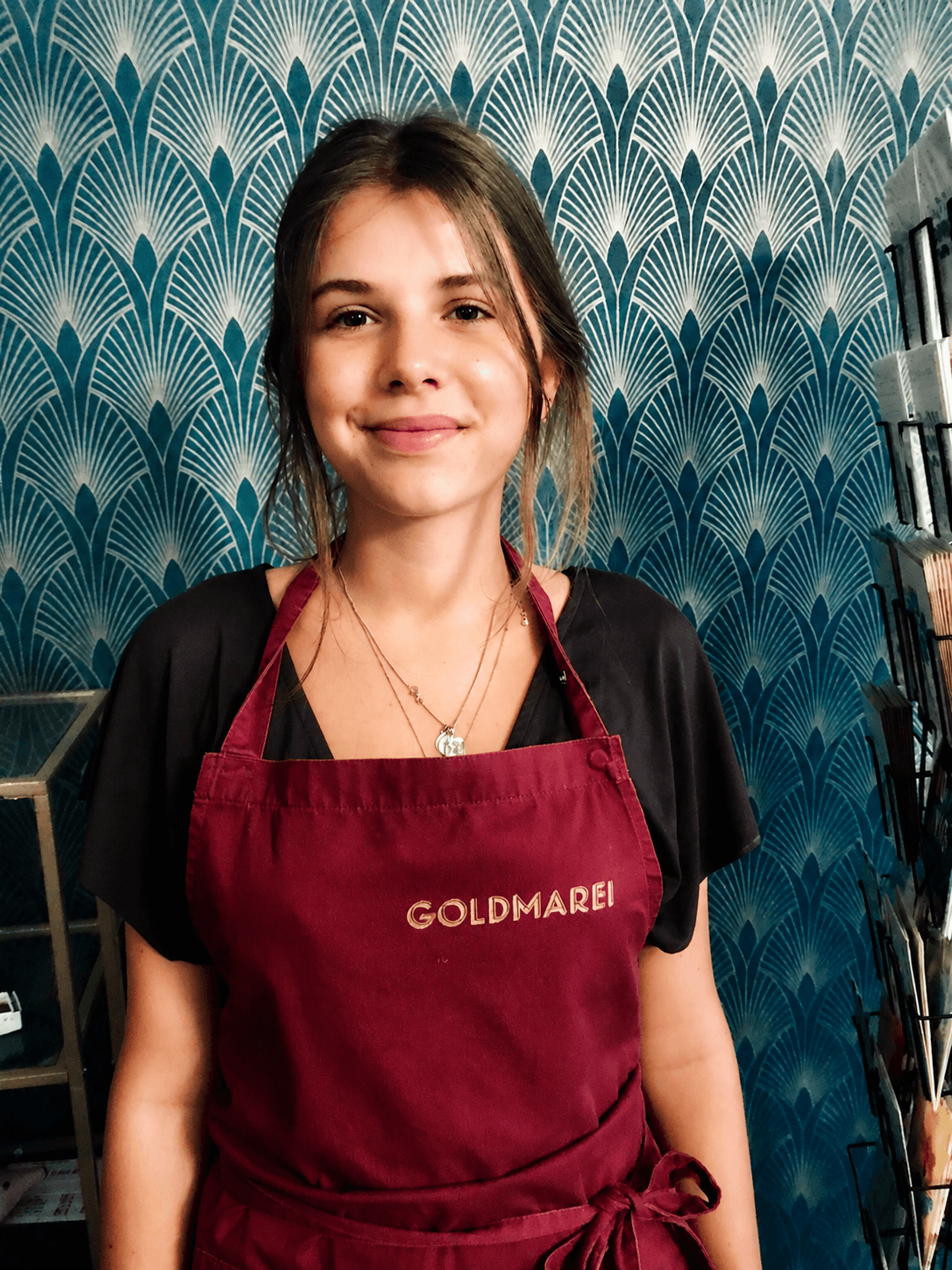 Junge Frau trägt eine rote Schürze mit der Aufschrift 'GOLDMAREI' in einem Geschäft oder Café, vor einer blauen, stilvollen Wand. Sie lächelt freundlich.