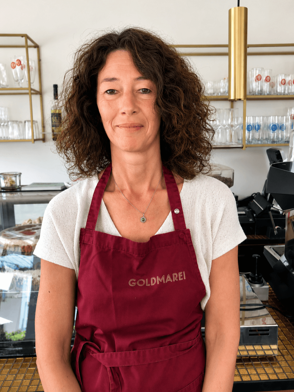 Frau trägt eine rote Schürze mit der Aufschrift 'GOLDMAREI' und steht in einer Kaffeebar oder Bäckerei.
