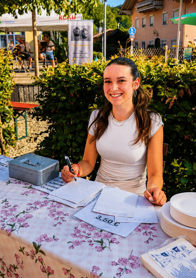 Eine lachende Frau sitzt an einem Tisch mit Blümchentischdecke vor einem Stand im Freien, schreibt auf Zettel und verkauft wahrscheinlich Lebensmittel oder Dienstleistungen, mit einem Preisschild von 3,50 €. Im Hintergrund sind andere Marktstände und Gebäude zu sehen.