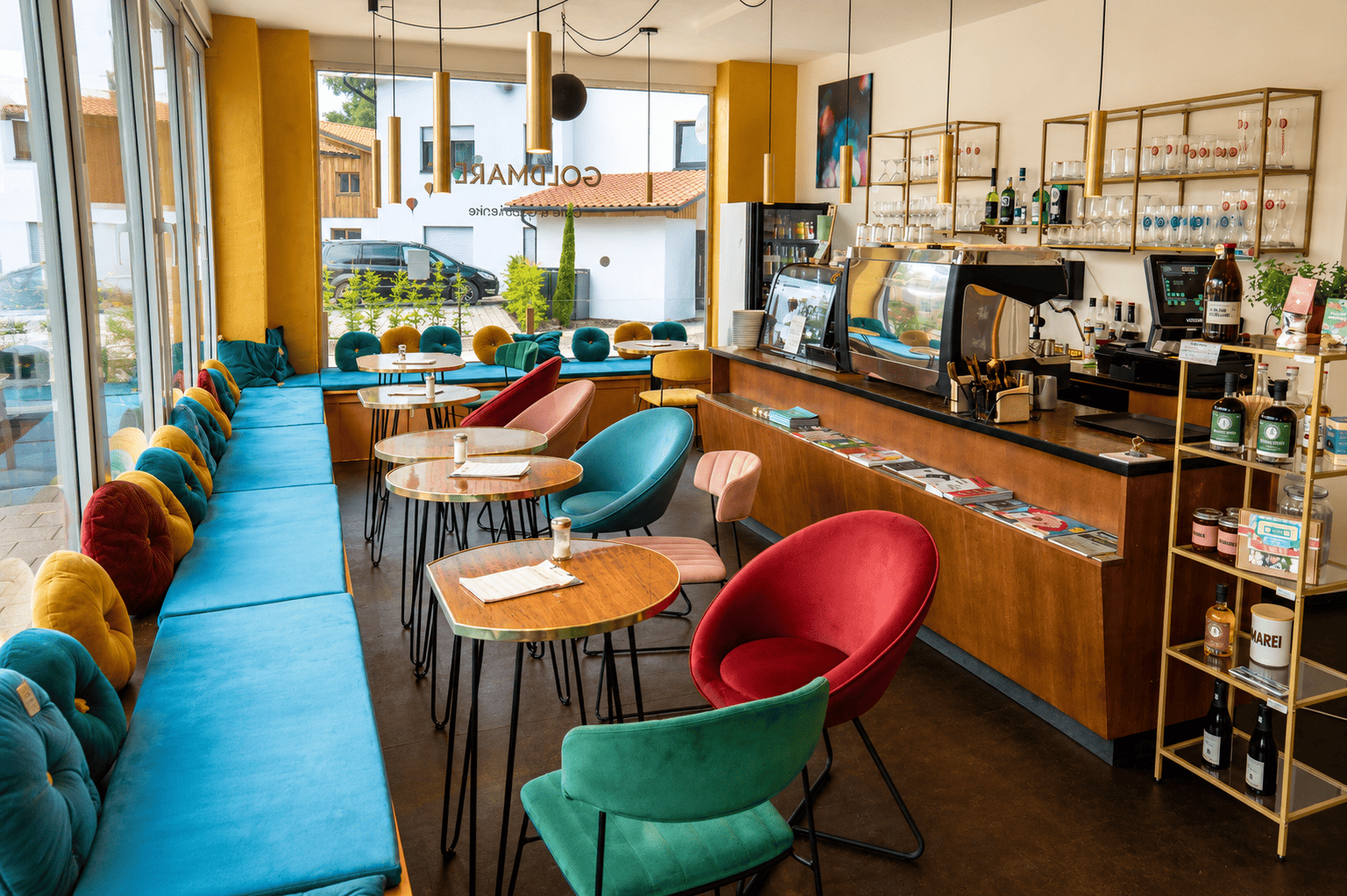 Innenaufnahme eines modernen Cafés mit bunten Sesseln und Tischen, Baristas an der Theke, große Fenster mit Blick nach draußen, dekorative Regale mit Gläsern und Flaschen, dabei gemütliche Atmosphäre.