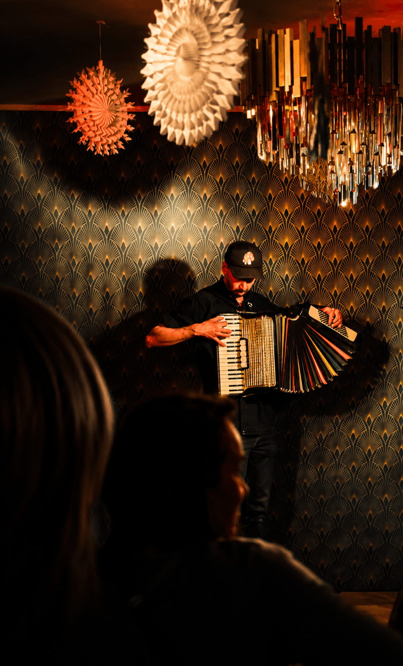 Ein Musiker spielt auf einem Akkordeon, während eine kleine Gruppe von Menschen im Dunkeln zusehen. Der Raum ist dekoriert mit schwarzen und goldenen Mustern an der Wand sowie schimmernden Kronleuchtern an der Decke.