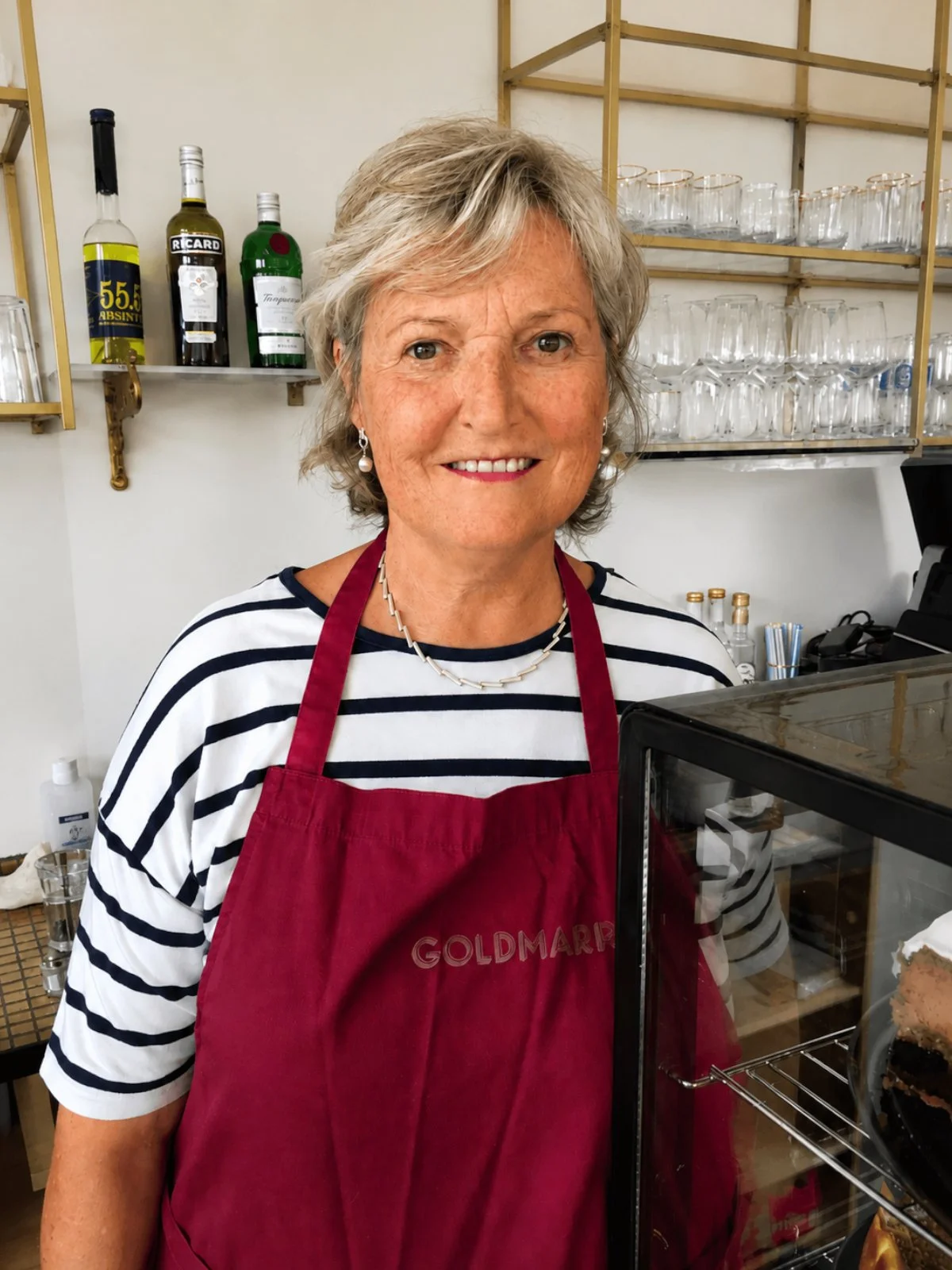 Eine ältere Frau trägt ein rotes Kochschürze und lächelt vor einer Bäckerei oder Kaffee. Sie steht neben einem Kuchenständer mit einem Stück Kuchen.