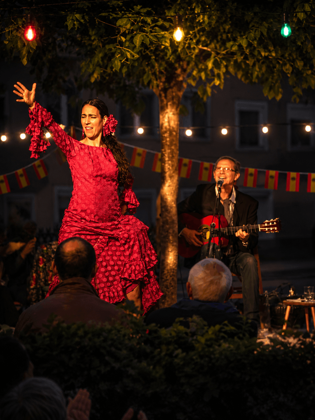 Feierabend im Freien mit einem Flamenco-Tänzerin in einem roten Kleid mit Pailletten, einem Gitarristen im Hintergrund, der singt und Gitarre spielt, bei abendlicher Beleuchtung und festlicher Dekoration.