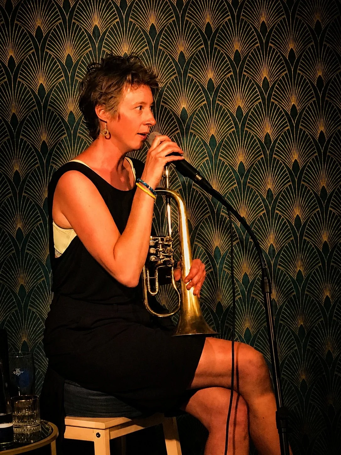 Frau spielt Trompete und singt in einem Restaurant mit goldfarbenem Art Deco Muster auf der Wand im Hintergrund.