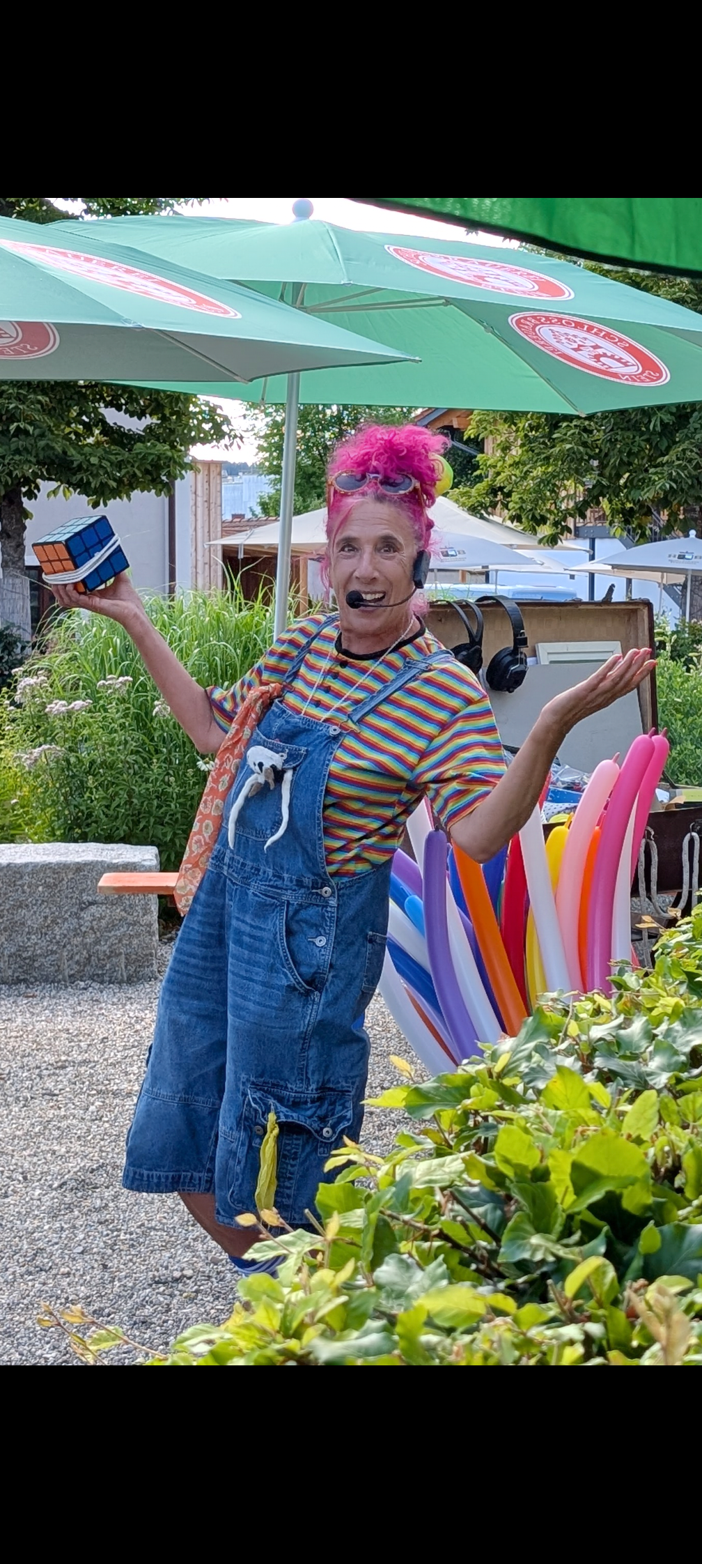 Eine fröhliche Frau mit pink gefärbtem Haar trägt bunte Kleidung und steht im Garten vor bunten Puppenstäben, umgeben von Sonnenschirmen und Pflanzen.