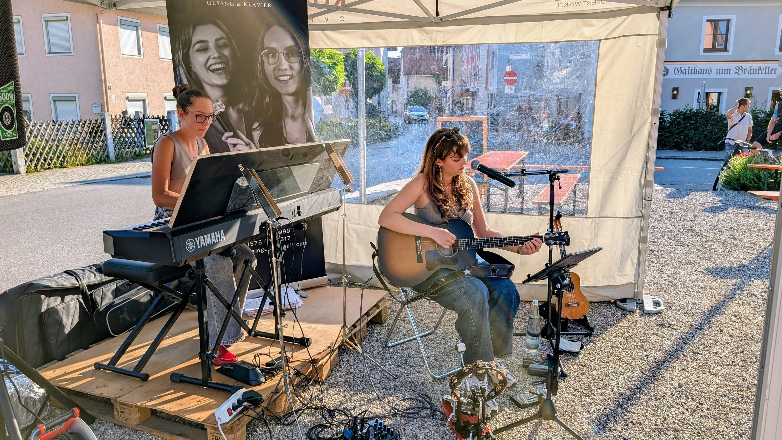 Zwei Frauen spielen Musik in einem Zelt im Freien. Eine spielt Klavier, die andere Gitarre. Im Hintergrund sind Häuser und Leute zu sehen.