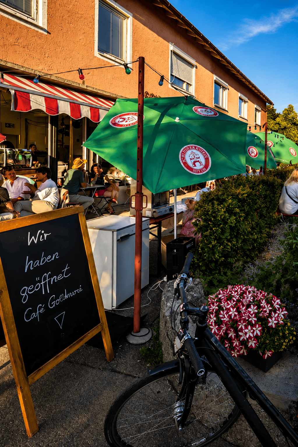 Ein Café im Freien mit mehreren grünen Sonnenschirmen, Blumen und Gästen, die zusammen sitzen, sowie ein Fahrrad im Vordergrund und ein schwarzes Brett mit der Aufschrift „Wir haben geöffnet Café Götzmeer“.