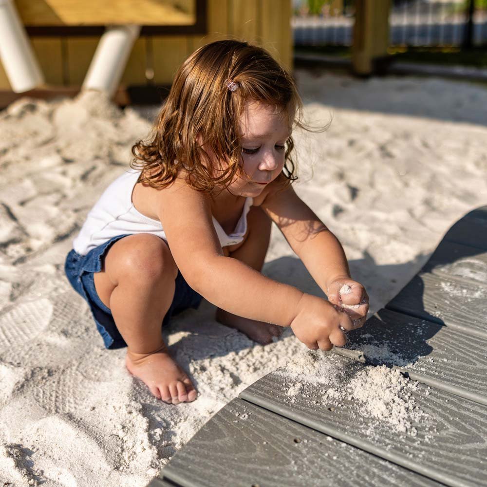 A young child with red hair and a white shirt playing in the sand on a beach or sandbox, scooping sand with her hands near a wooden board or deck.