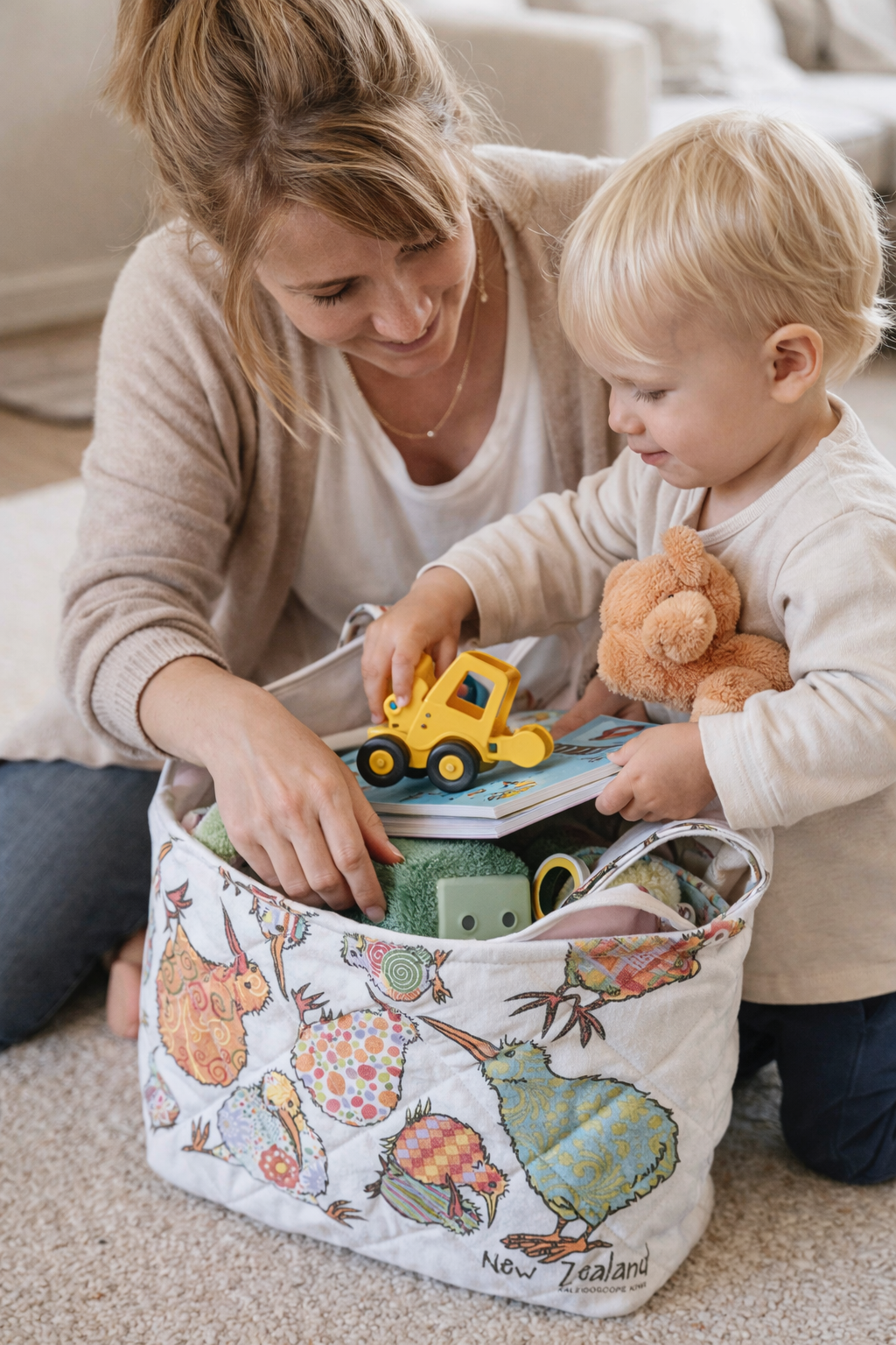A woman and a young boy are sitting on the carpeted floor, going through a large tote bag filled with toys and books. The boy holds a teddy bear and places a yellow toy bulldozer on top of a book, smiling. The tote bag has colorful bird illustrations and says 'New Zealand' on it.