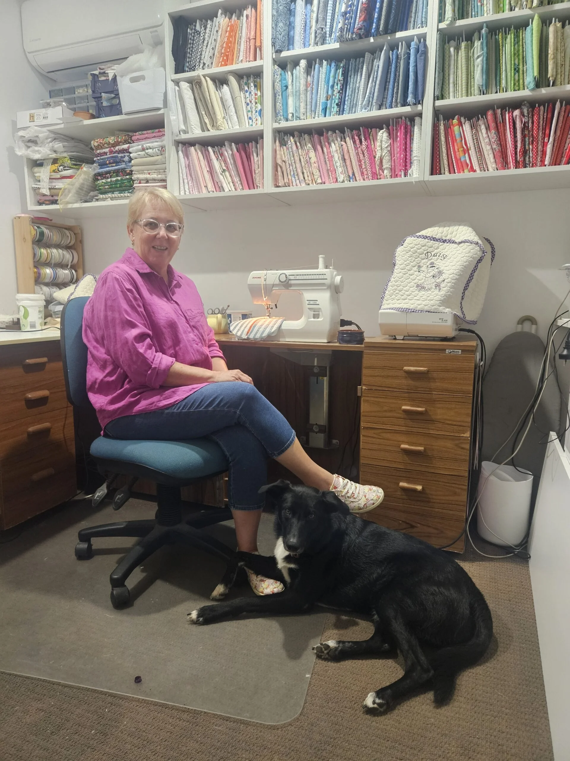A woman sitting in a sewing room with a black dog lying on the floor beside her. She is smiling and wearing a pink shirt and jeans. The room has a sewing machine on the desk and shelves filled with colorful fabric.