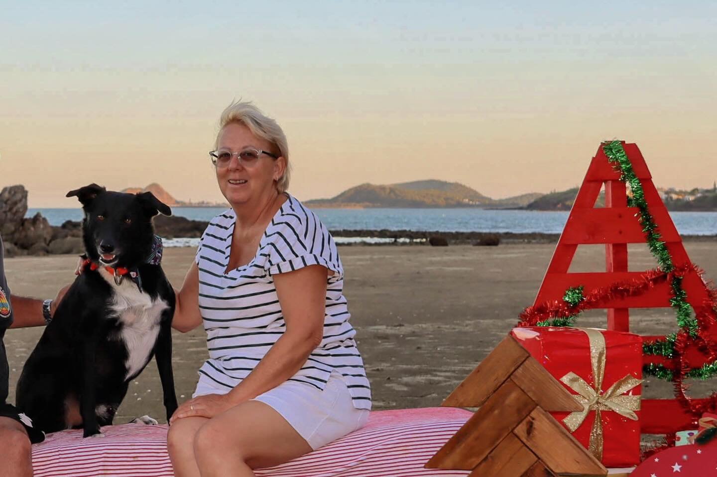 A woman with short blonde hair, glasses, and a striped shirt, sitting on a pink striped blanket on a beach, smiling with a black and white dog beside her. There are holiday decorations, including a red wooden Christmas tree and presents, in the foreground, with the ocean and distant islands in the background.