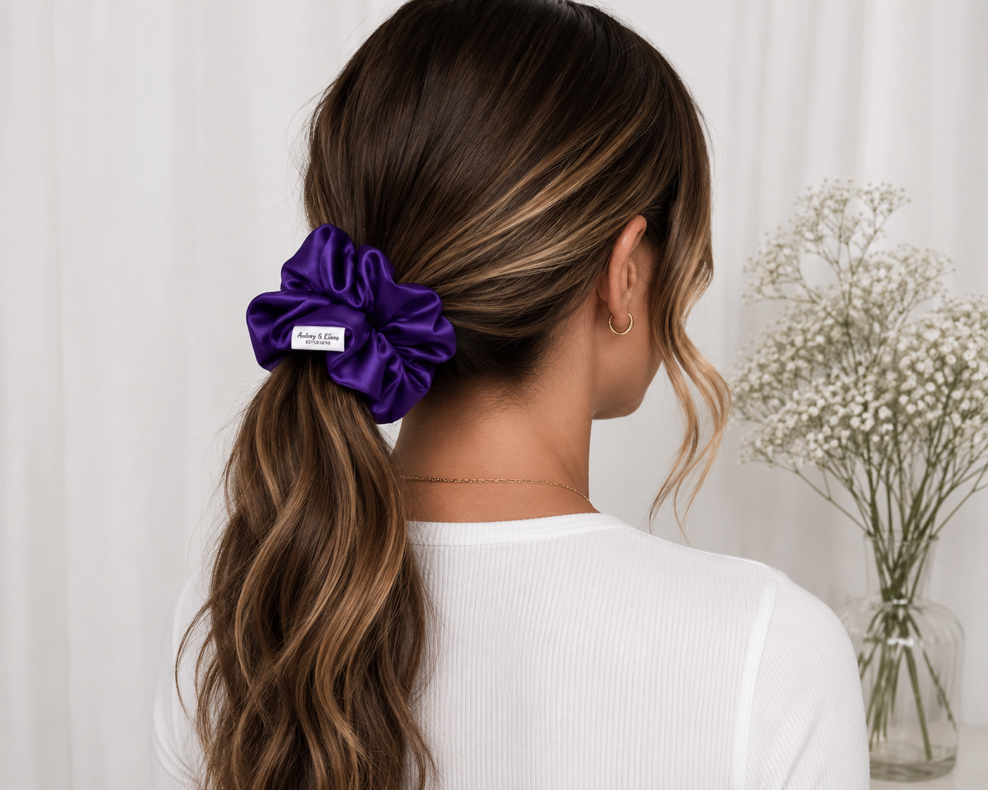 Back view of a woman with wavy brown hair in a ponytail secured with a purple scrunchie, wearing a white top and small hoop earrings, with white flowers in a clear vase in the background.